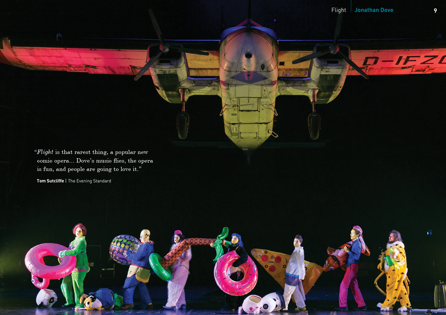 An aircraft hangs upside down from the ceiling above a stage with performers in colorful costumes holding inflatable objects, with a dark background.