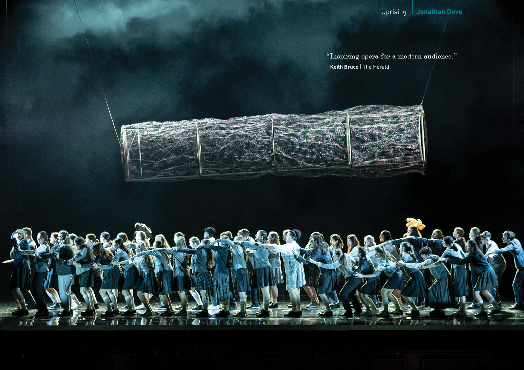 Stage performance with numerous students in uniforms, pulling a long rope, and a large floating rectangular structure above them, with dark dramatic clouds in the background.