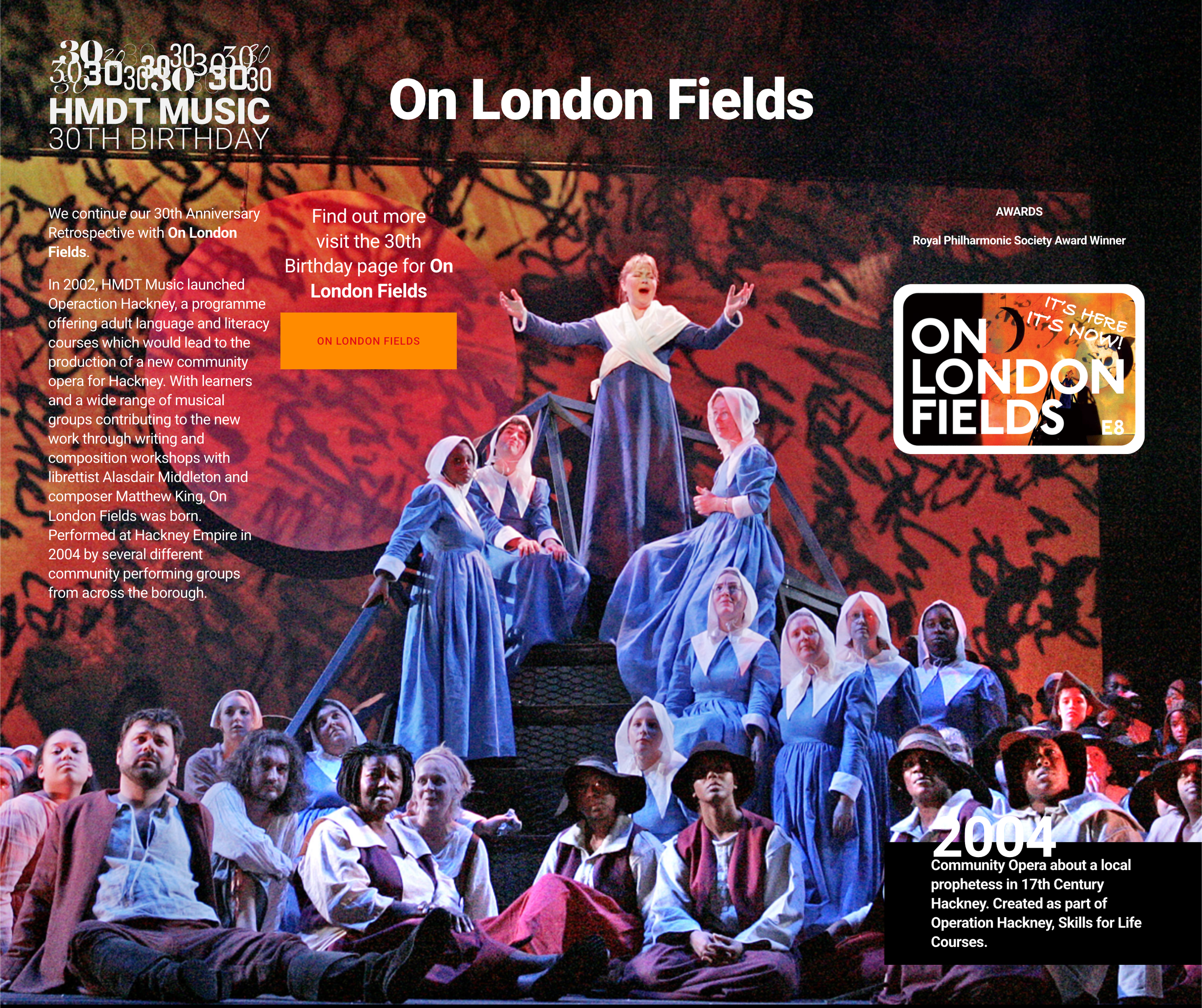 Scene from a community opera performance with numerous actors dressed in period costumes, set on a stage with a dark background, featuring a woman in a long blue dress with a white shawl center stage, surrounded by others in similar costumes.