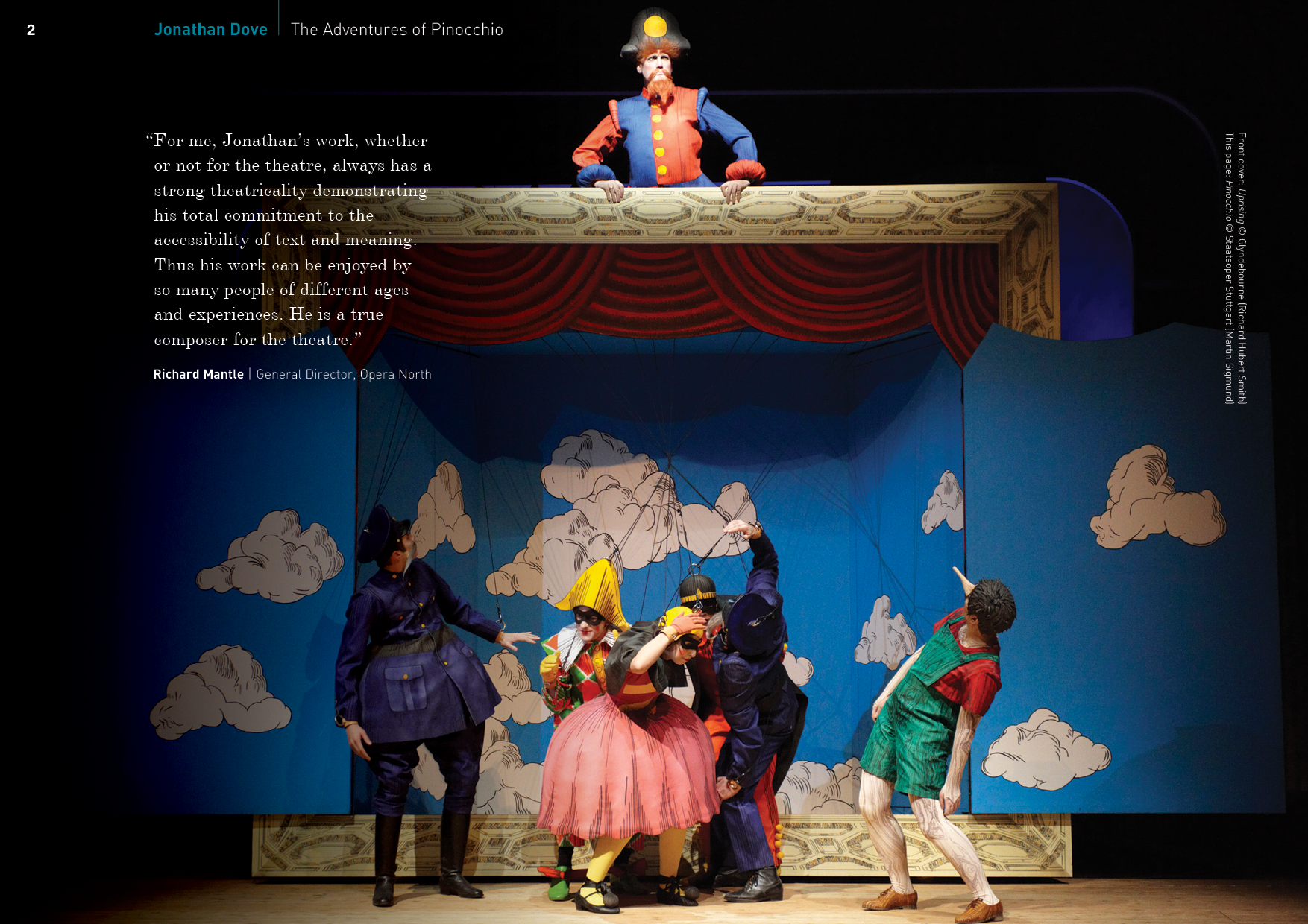 A theatrical stage scene from Pinocchio with puppeteers operating marionettes against a cloud-painted blue backdrop, with a person in costume above the stage observing.