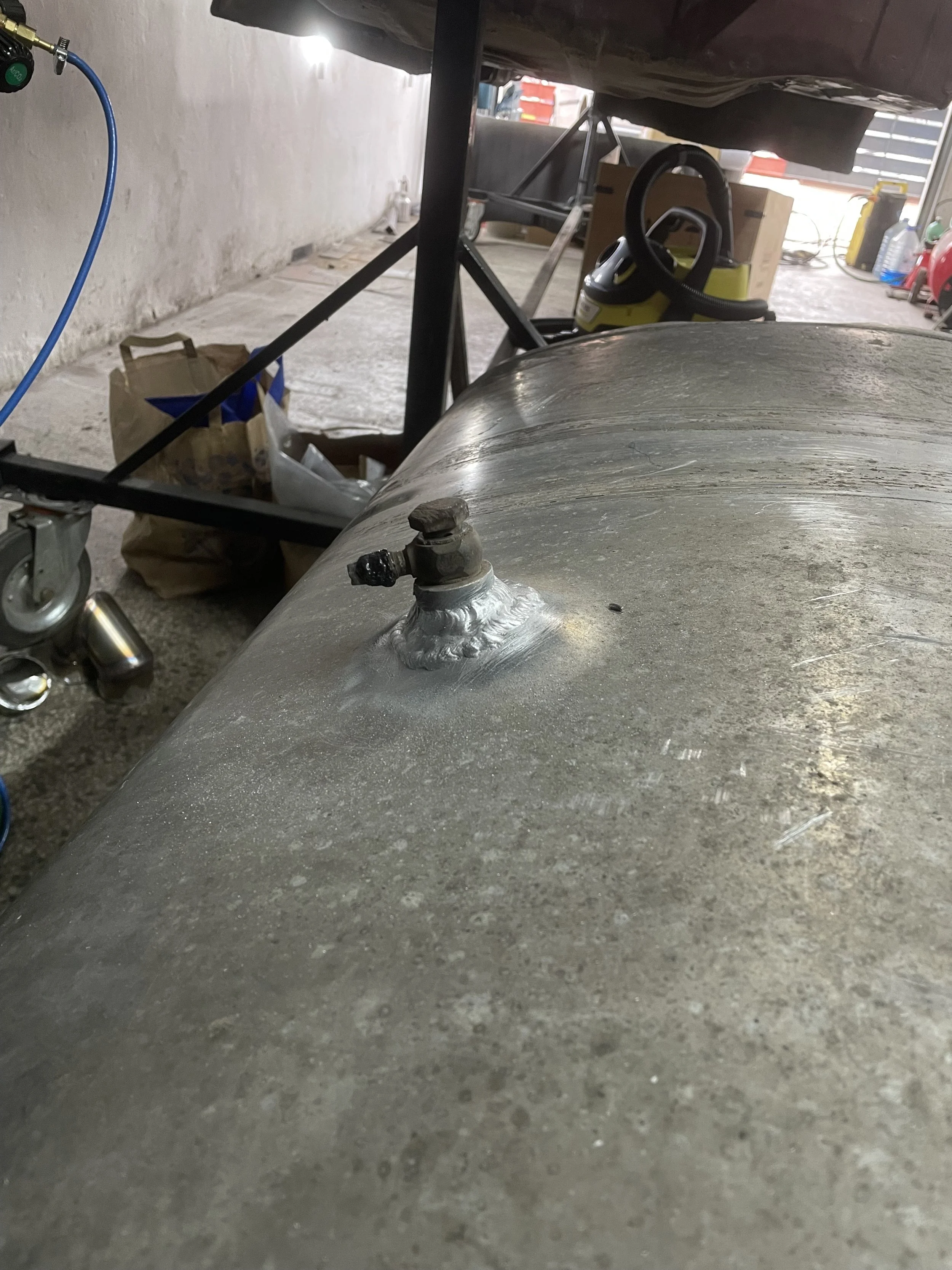 Close-up of a metallic surface with a welded fixture and a valve protruding from it, in a workshop or garage setting.