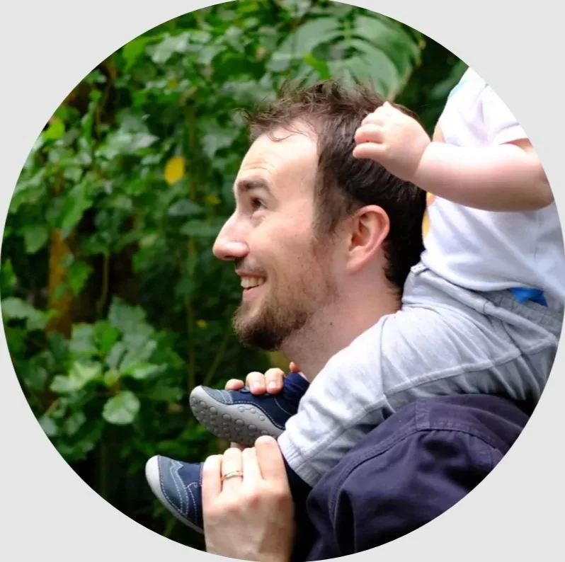 A man with a beard and short hair carrying a young child on his shoulders outdoors with green foliage in the background.