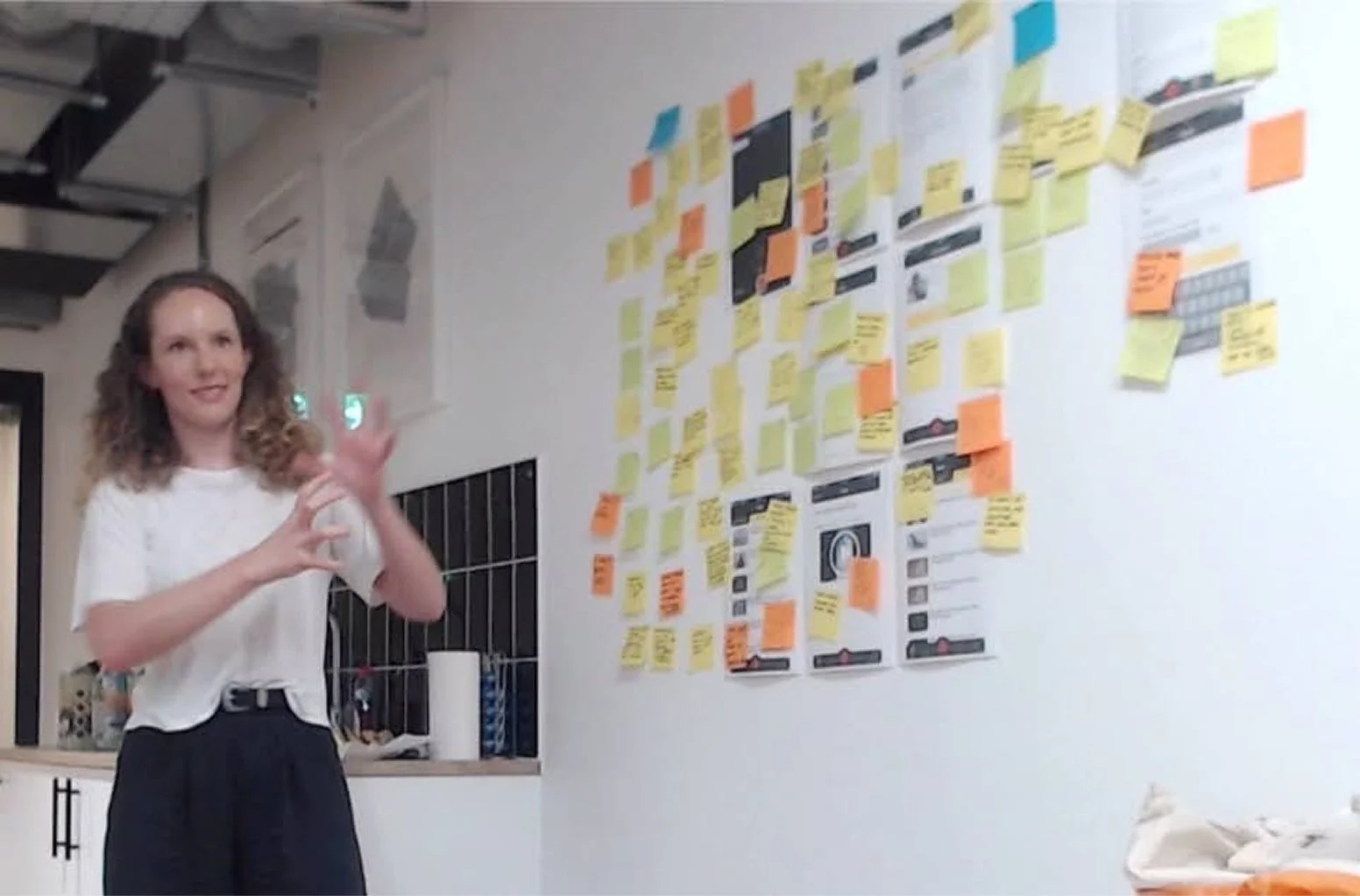 Interior room with a woman standing next to a wall covered in colorful sticky notes and printed papers, some arranged in a grid pattern. She is smiling and making a hand gesture. There are kitchen items on a counter and a staircase visible in the background.
