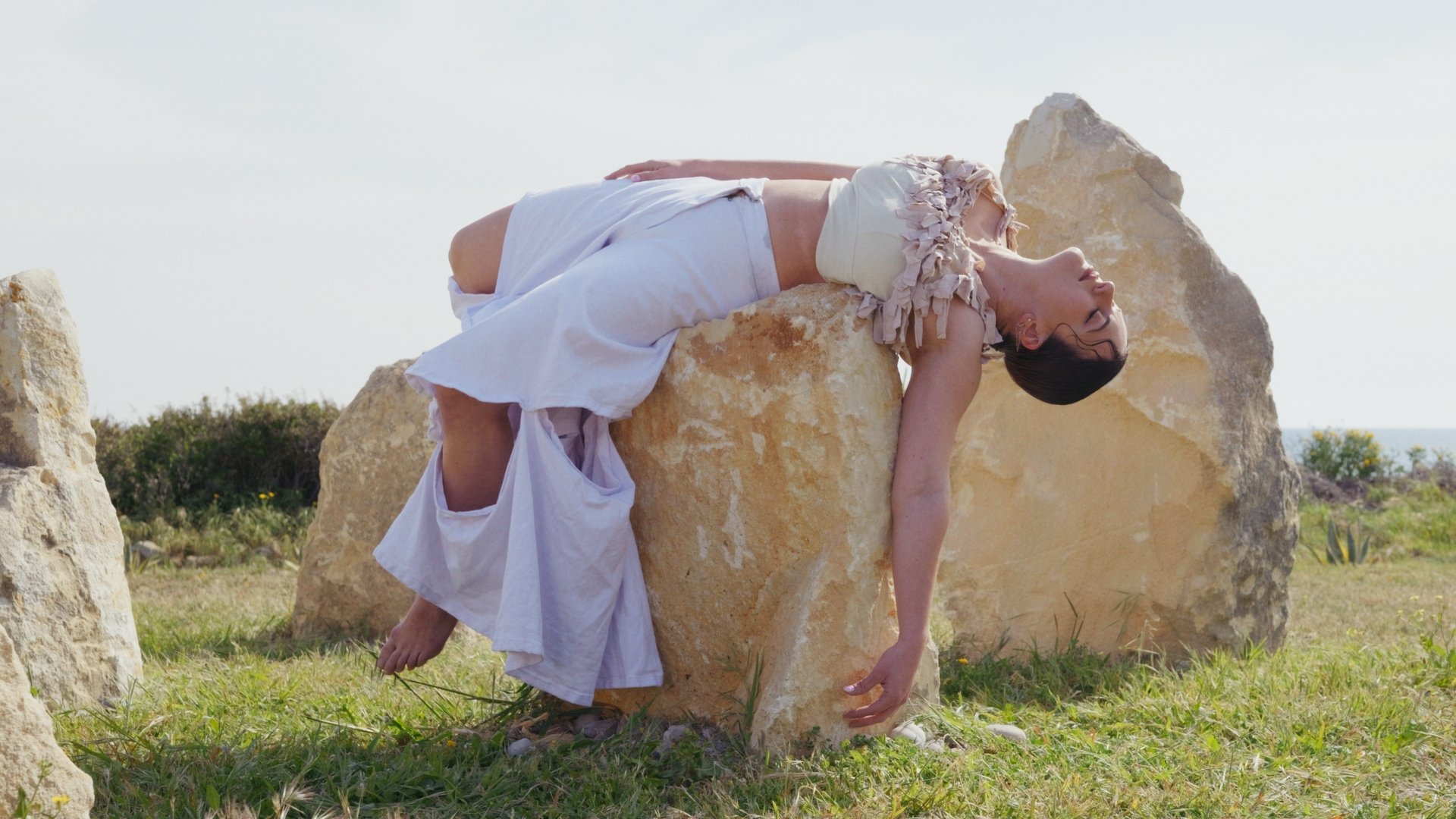 Person in white clothing reclined on large stone outdoors, with grassy landscape in background.