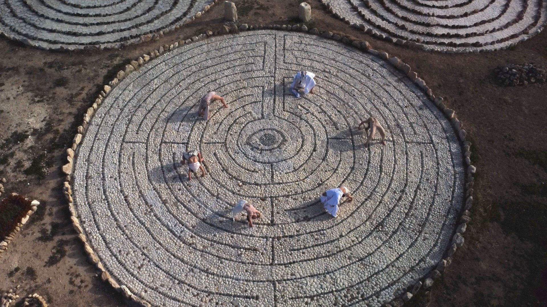 Aerial view of people in a circular stone labyrinth, performing dance or yoga on cobblestones.