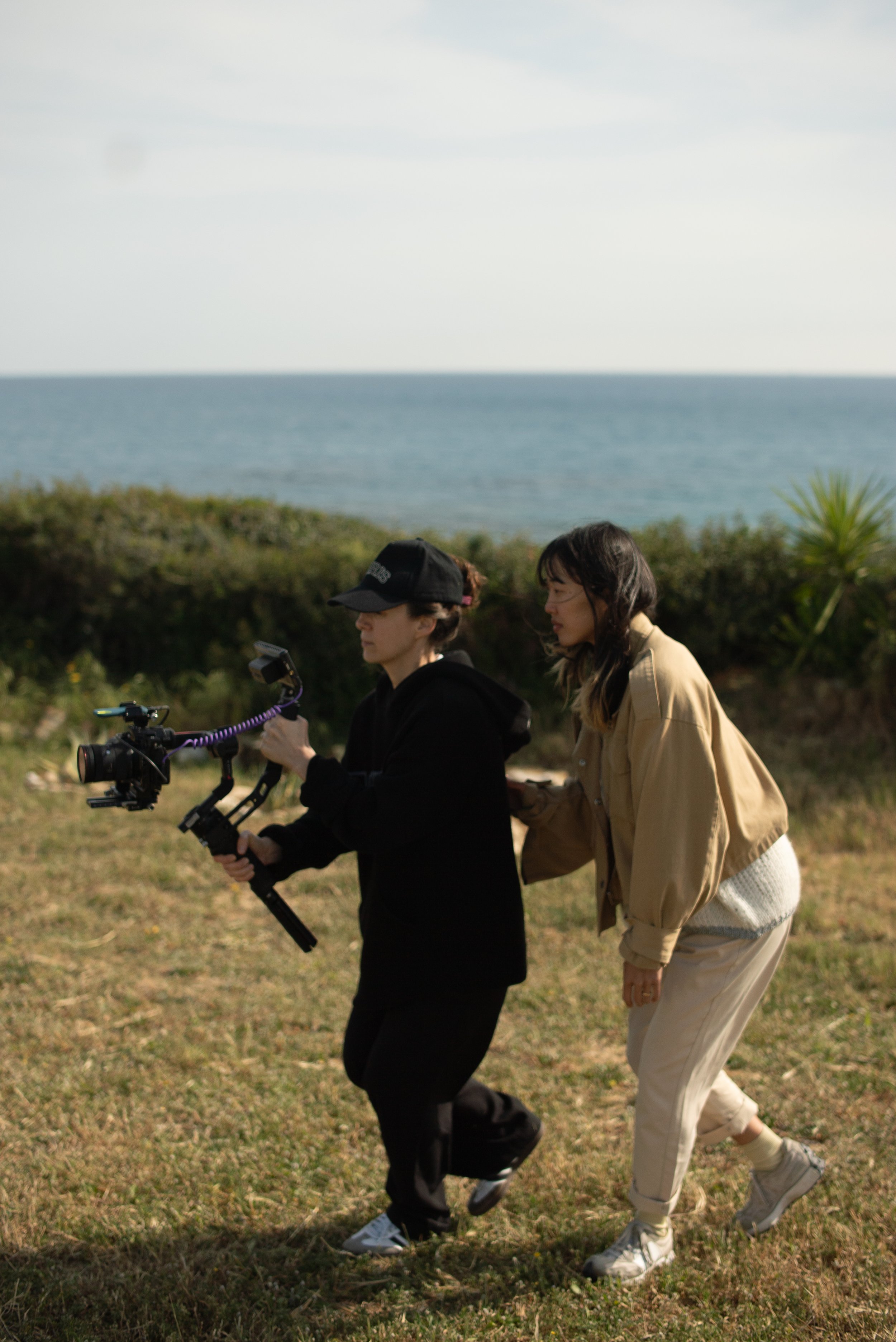 Two people outdoors near a coastline, one holding a camera stabilizer, the other assisting.