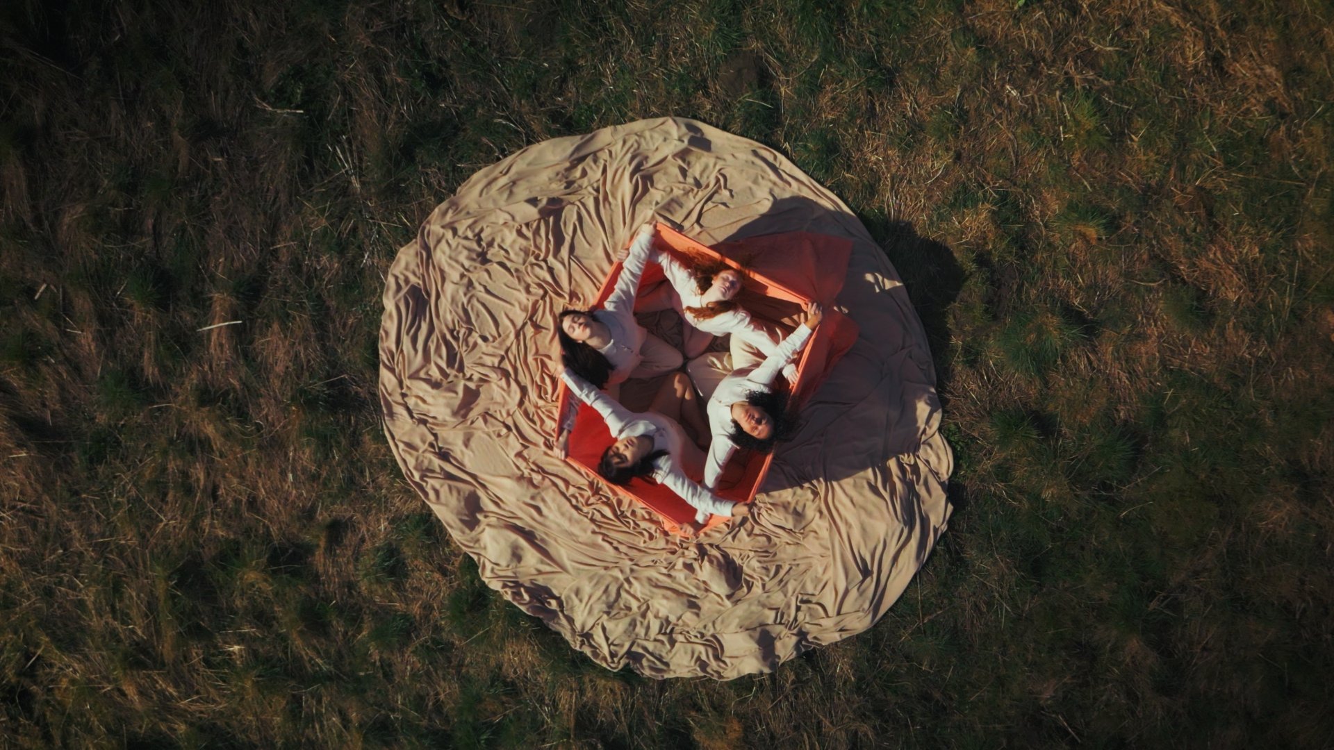 People lying on a red hammock surrounded by a large beige fabric on grass, viewed from above.