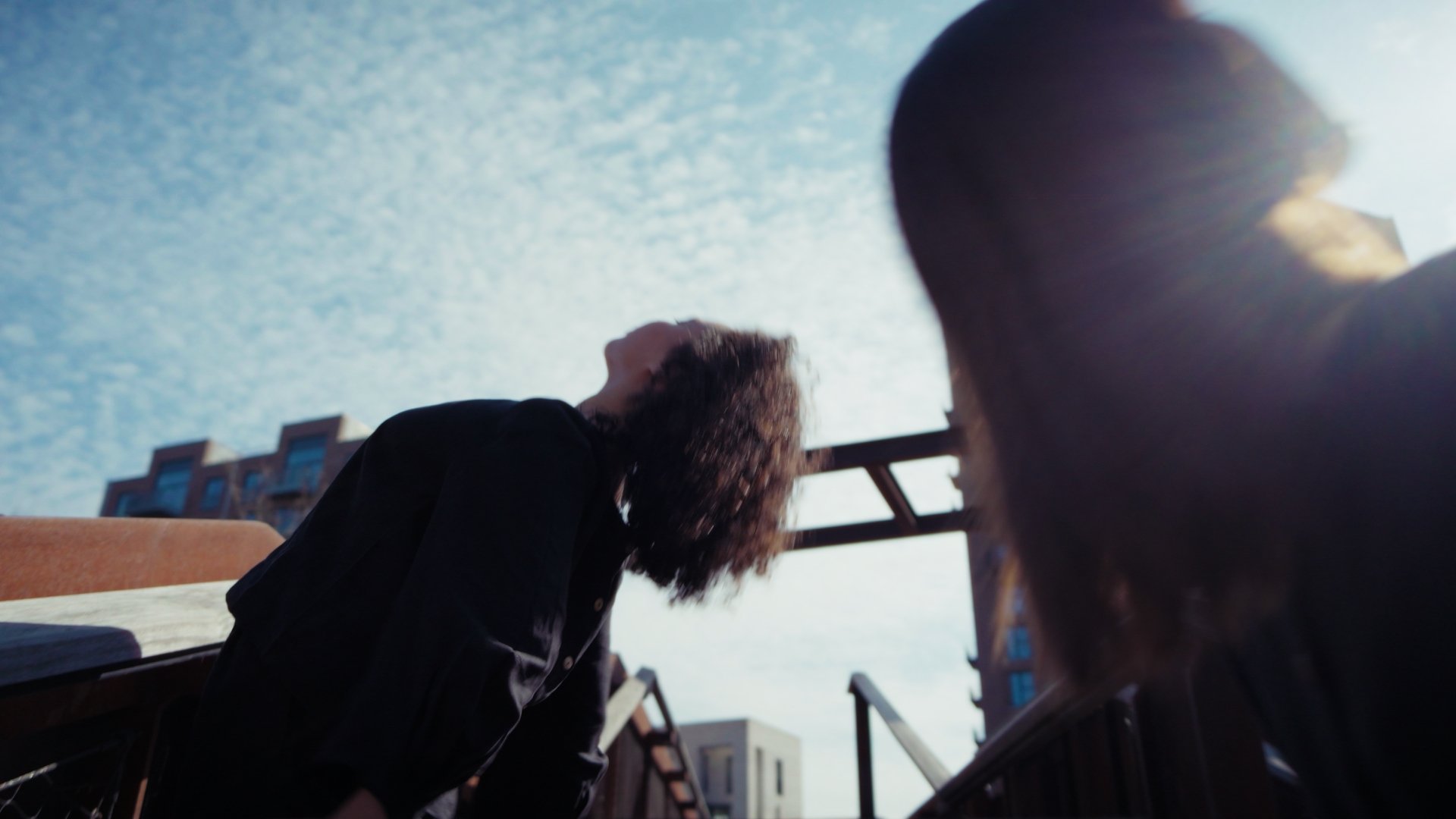 Two people outside under a blue sky with clouds, looking upwards. One person is in shadow on the right, while the other has curly hair and wears a black jacket. Urban buildings and a metal structure are visible in the background.