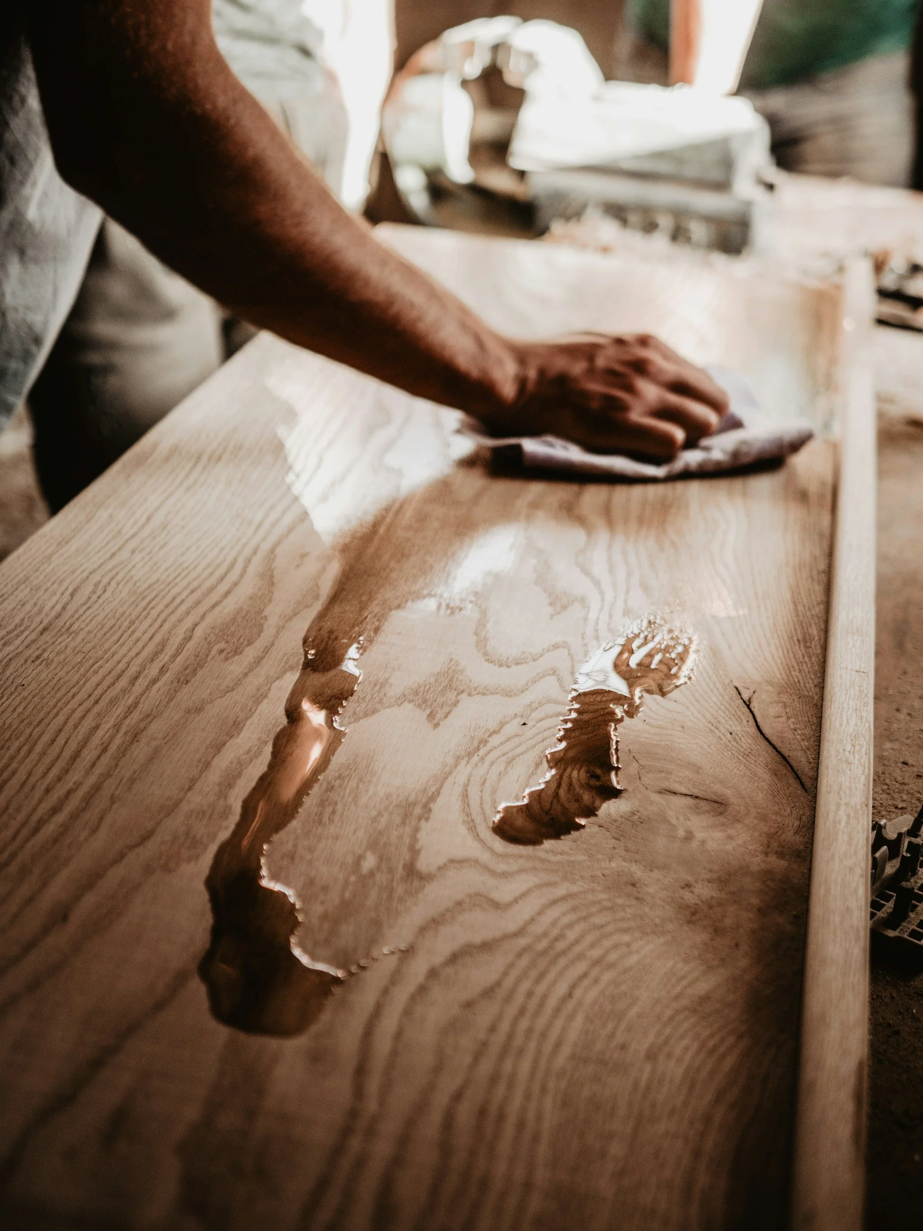 A person is sanding or polishing a long wooden board in a woodworking workshop, with tools and wood pieces visible in the background.
