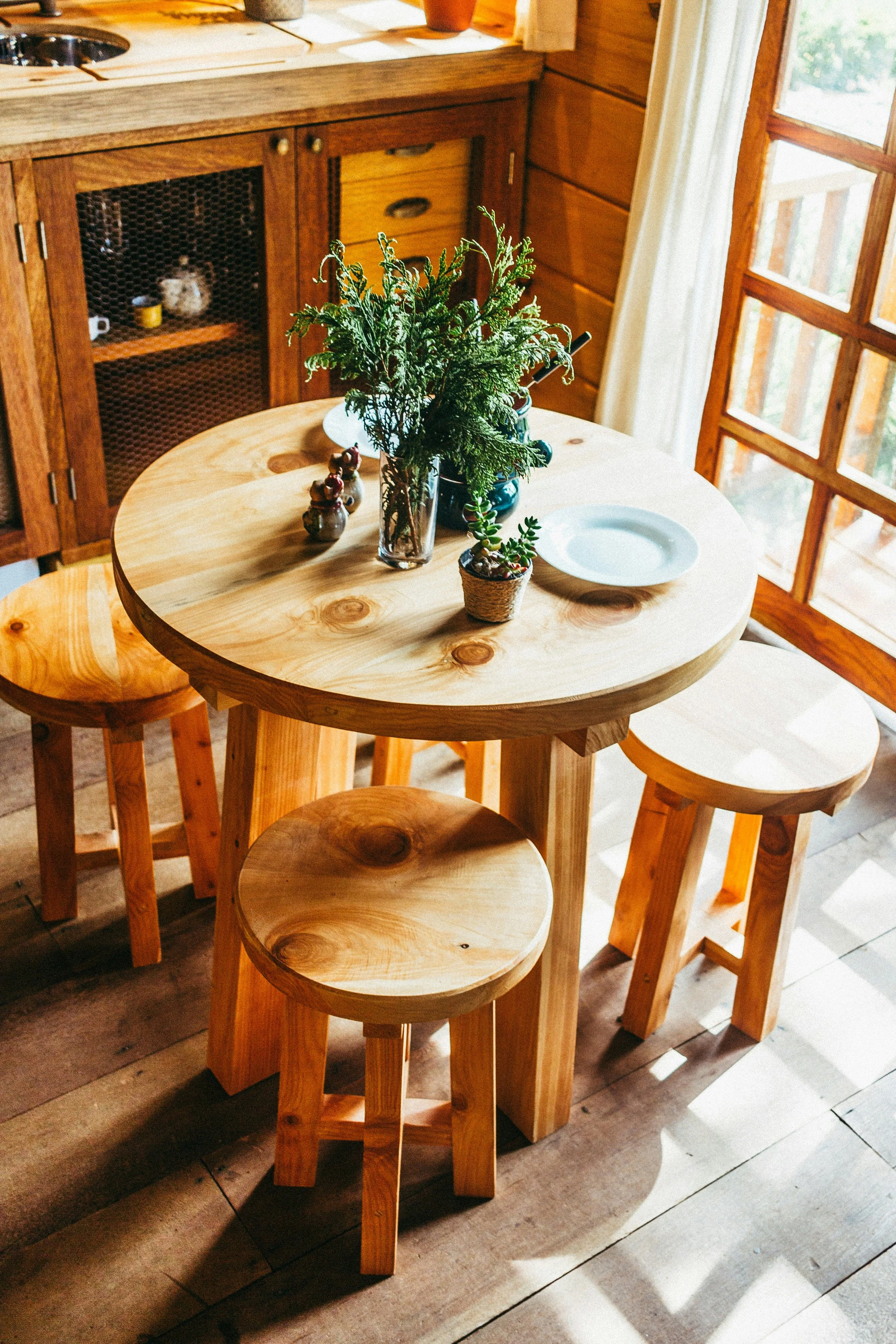 A wooden dining table with three matching stools, decorated with a pitcher of greenery, a small potted plant, and some figurines, in a cozy, sunlit kitchen with rustic wood cabinetry and a window with curtains.