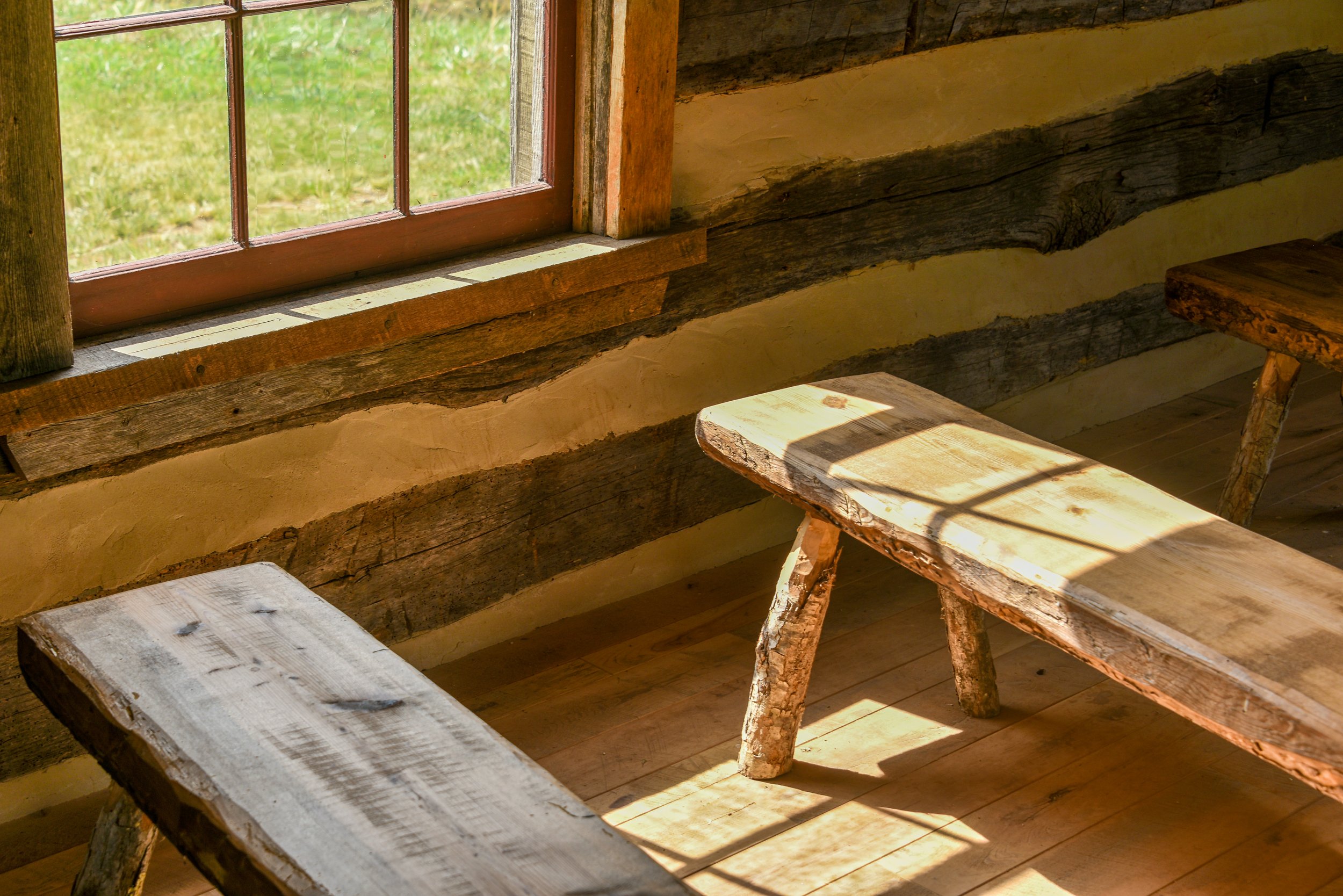 Interior of a rustic cabin with sunlight casting shadows on a wooden table and benches near a window showing a green outdoor scene.