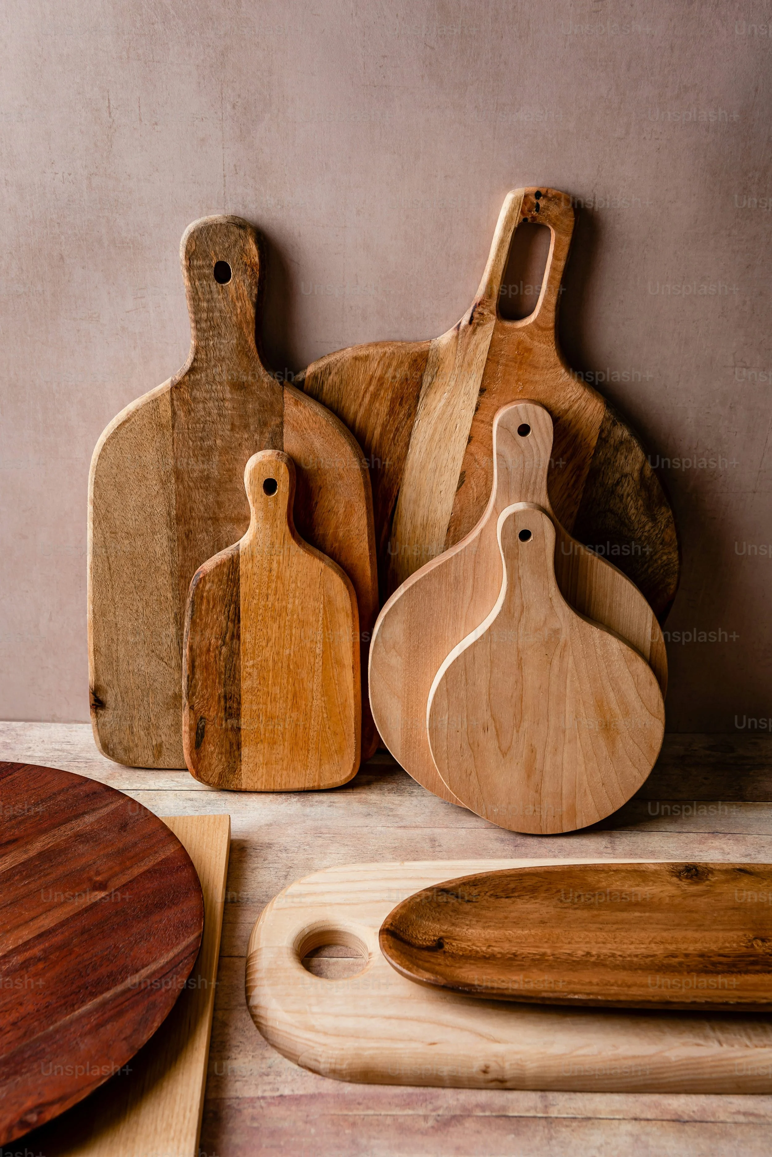 Several wooden cutting boards of various shapes and sizes, some with handle holes, are arranged upright against a pink wall. Additional wooden and ceramic dishes are partially visible in the foreground.