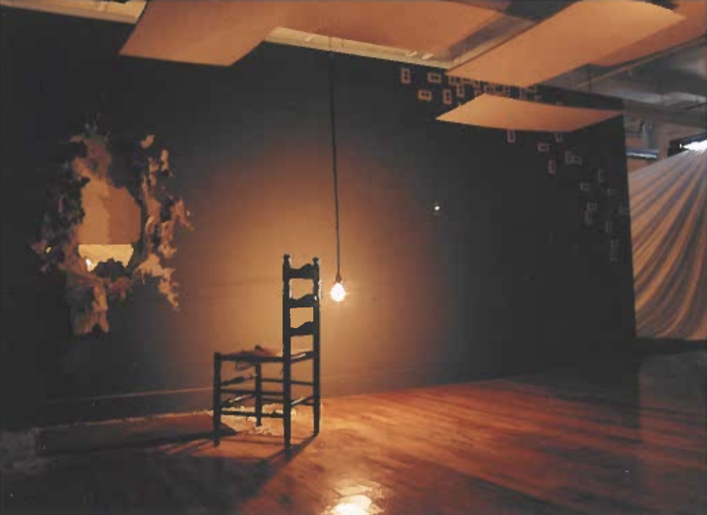 Empty wooden chair illuminated by hanging light bulb in a dimly lit room with dark walls, a small opening showing foliage, and colorful fabric draping on the right.