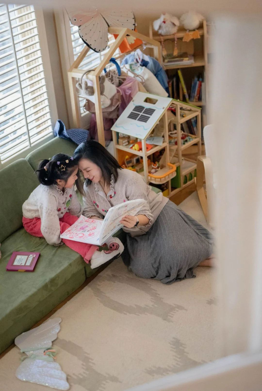 A woman and a young girl sitting on the floor of a cozy room, looking at a book together and smiling. The room has a green couch, a wooden bookshelf with toys and stuffed animals, and a window with blinds.