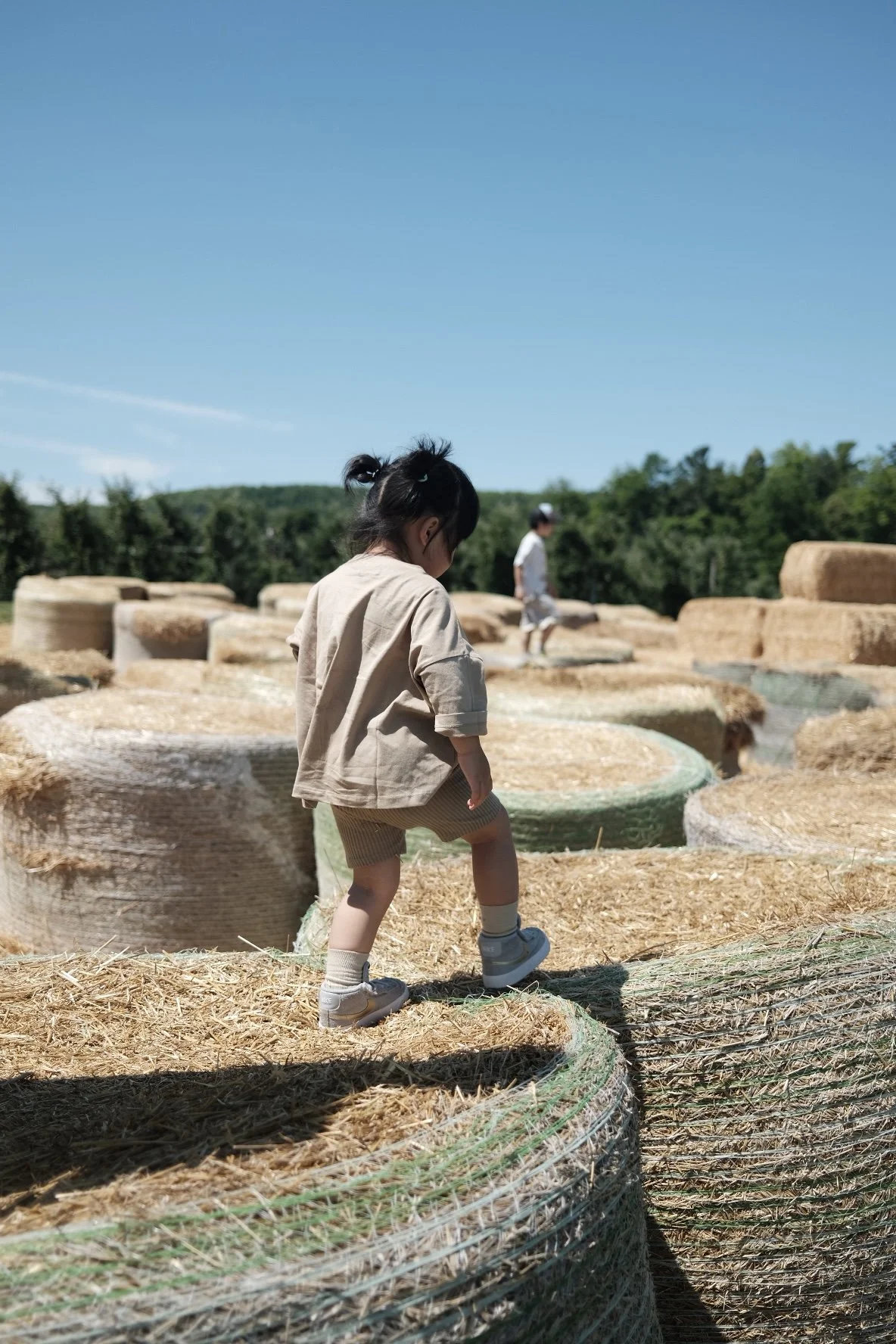 A child with dark hair tied in a ponytail, wearing a beige oversized shirt, shorts, and sneakers, stepping on hay bales outdoors on a sunny day. Another child is visible in the background among more hay bales, with a green, wooded landscape and blue sky behind.