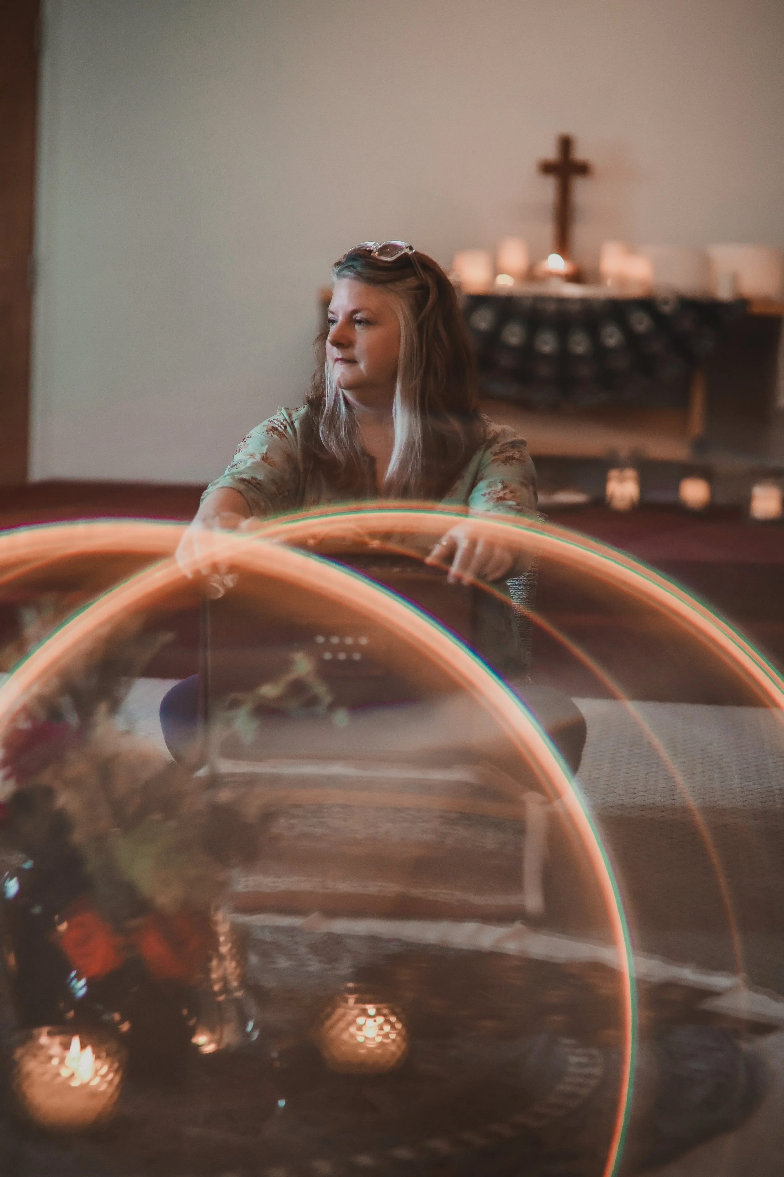 A woman with long, wavy hair sitting at a table with a blurred background of candles and a cross on the wall. The foreground has light trails creating arcs of glowing light.