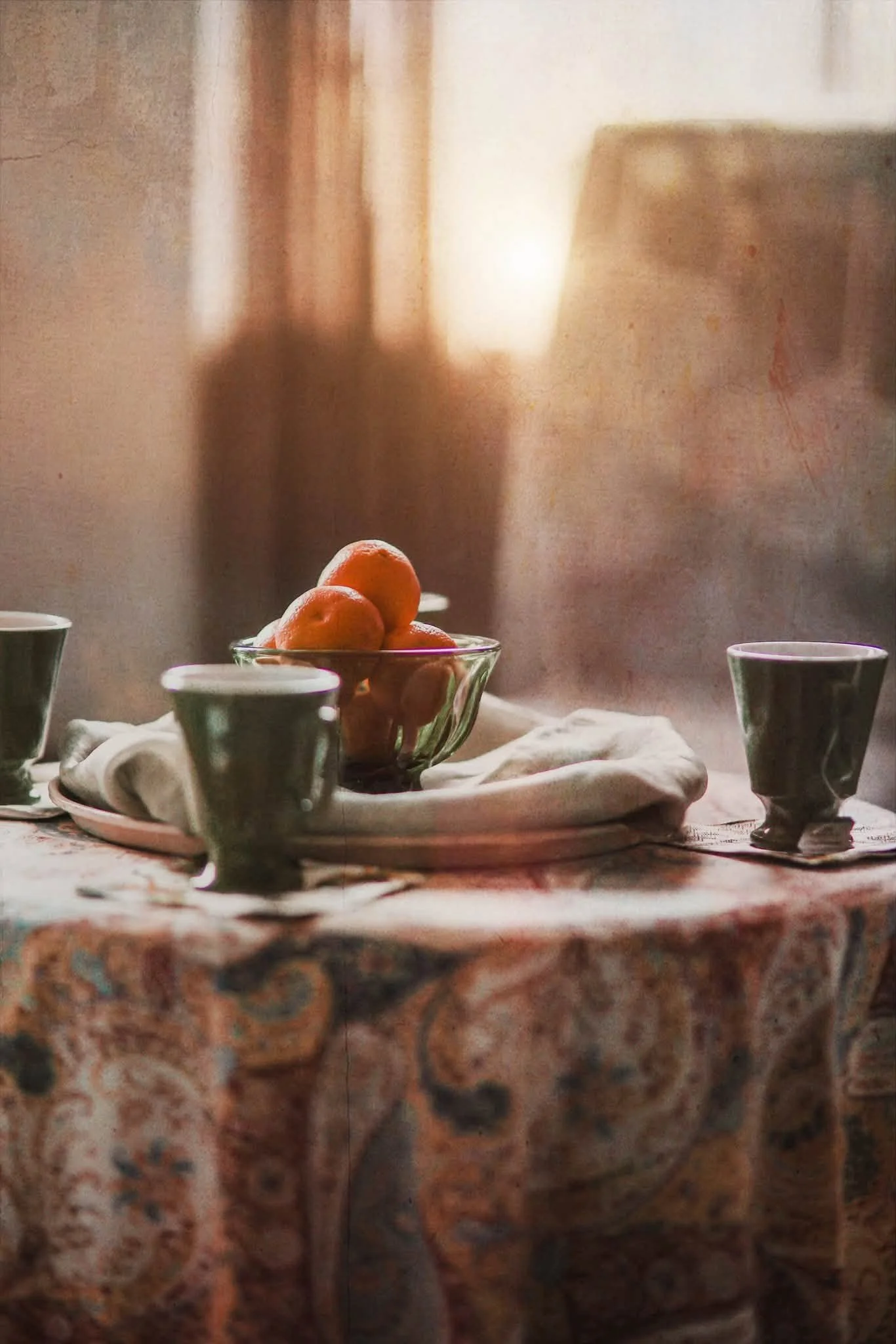 A table set with a white cloth, a glass bowl with oranges, surrounded by three cups, with a blurred background of a room with warm lighting.