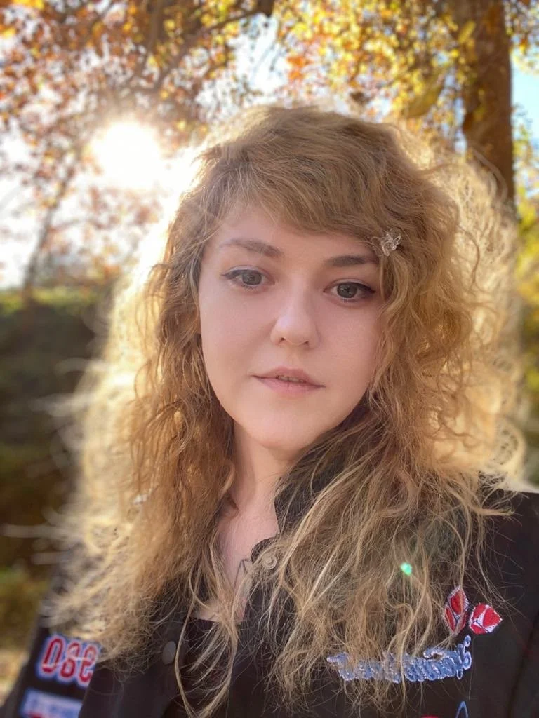 Person with curly hair smiling outdoors with sunlight and trees in the background.