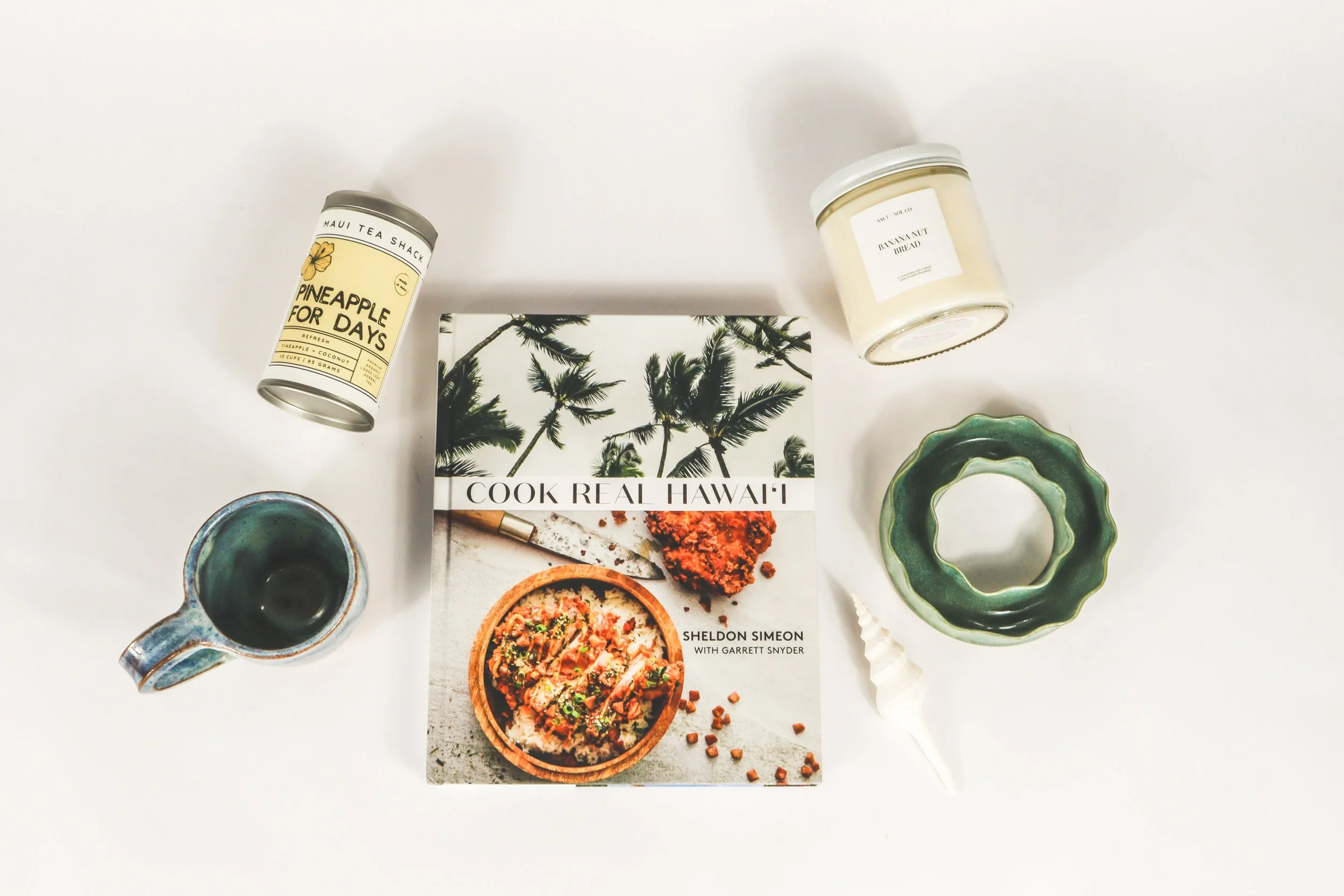 Hawaiian gift products laid out on a white background, including "Cook Real Hawaii" cookbook, handcrafted coffee mug, pineapple tea, banana nut bread candle, and ceramic lei making vase