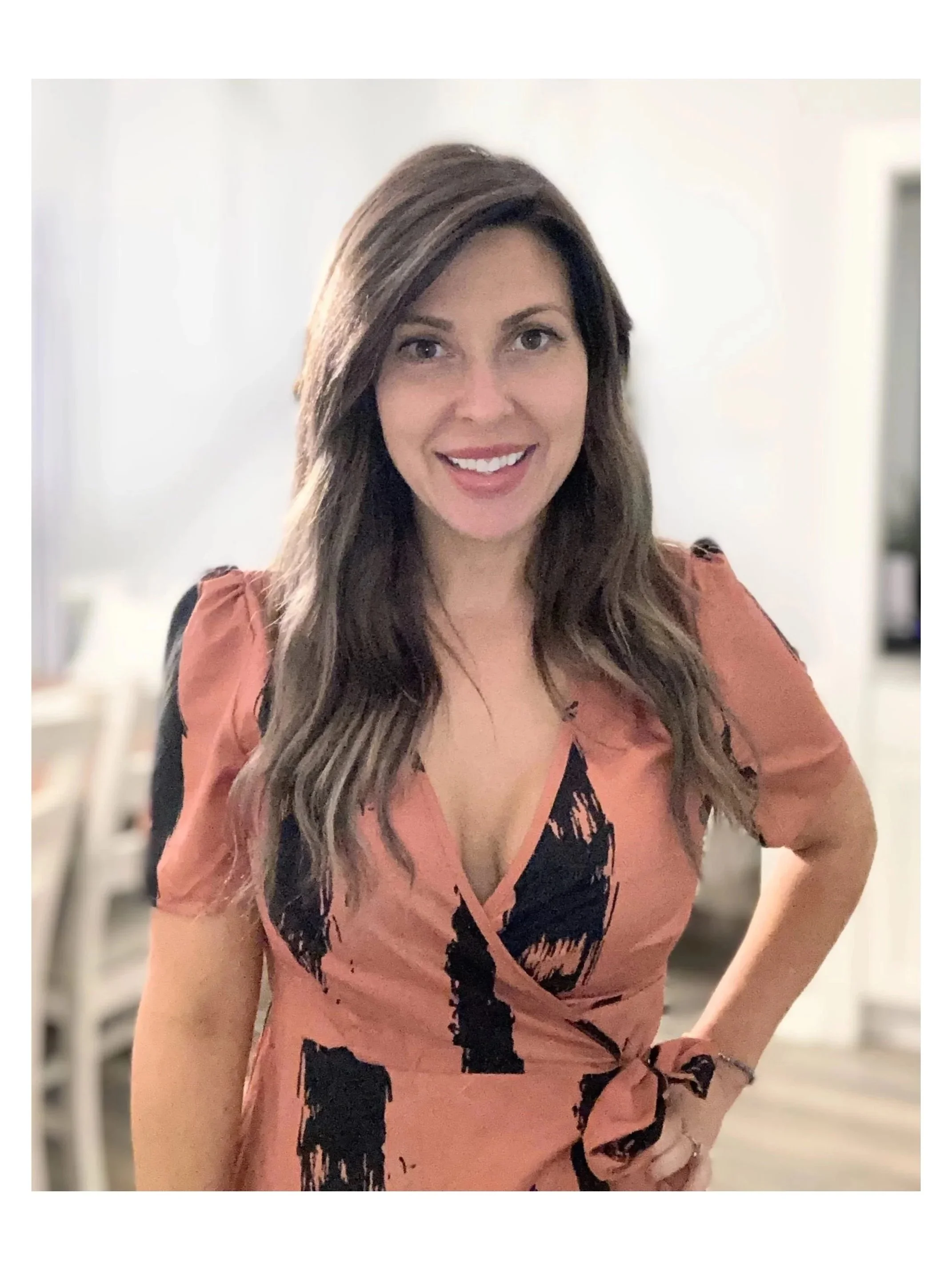 A woman with long, wavy brown hair, smiling with teeth showing, wearing a pinkish-orange dress with black pattern and a black, lace-up detail in the front. She is standing in a room with light-colored walls and furniture in the background.