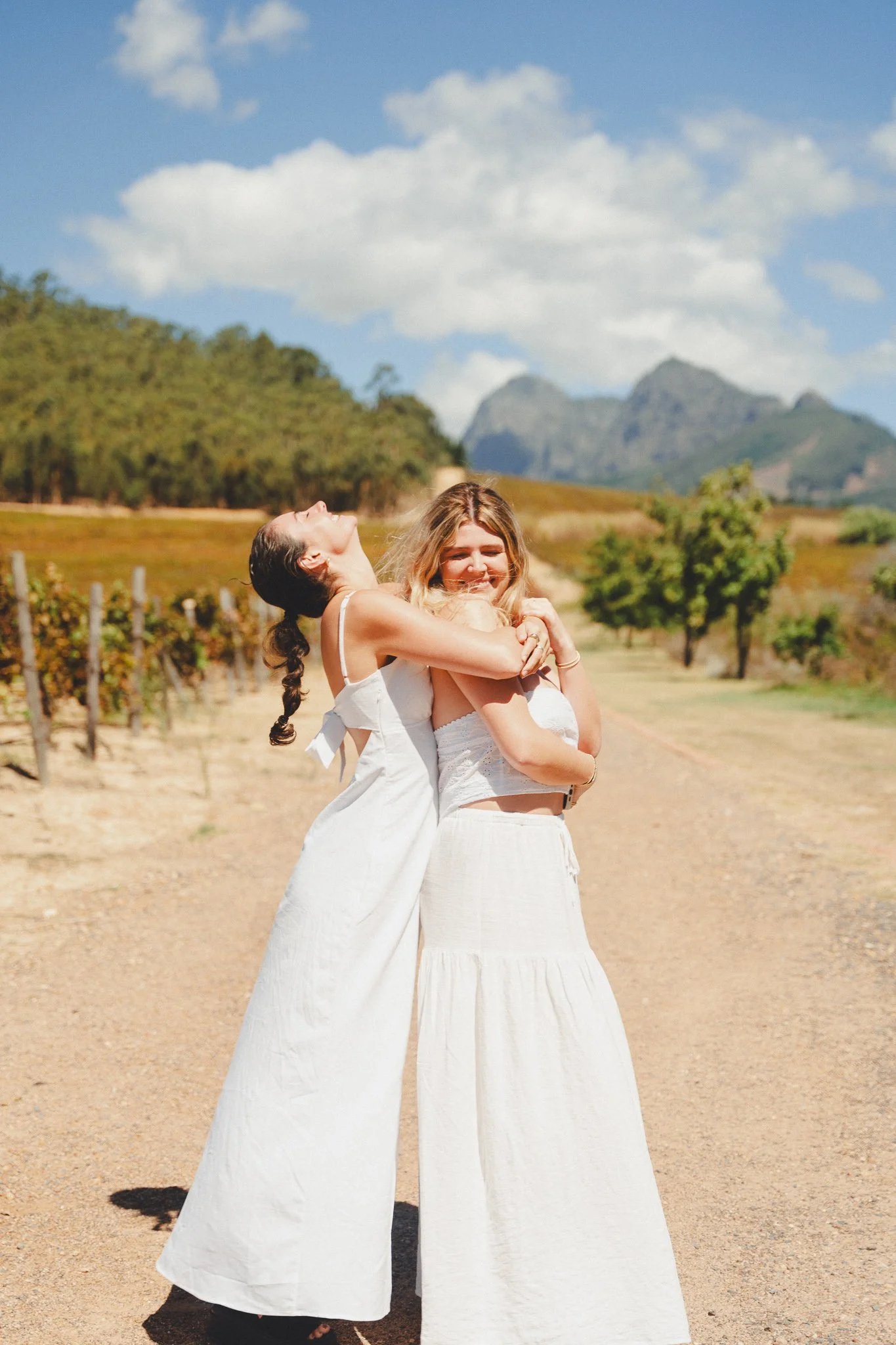 Two women hugging and laughing on a dirt path in a scenic outdoor vineyard setting with mountains in the background on a sunny day.