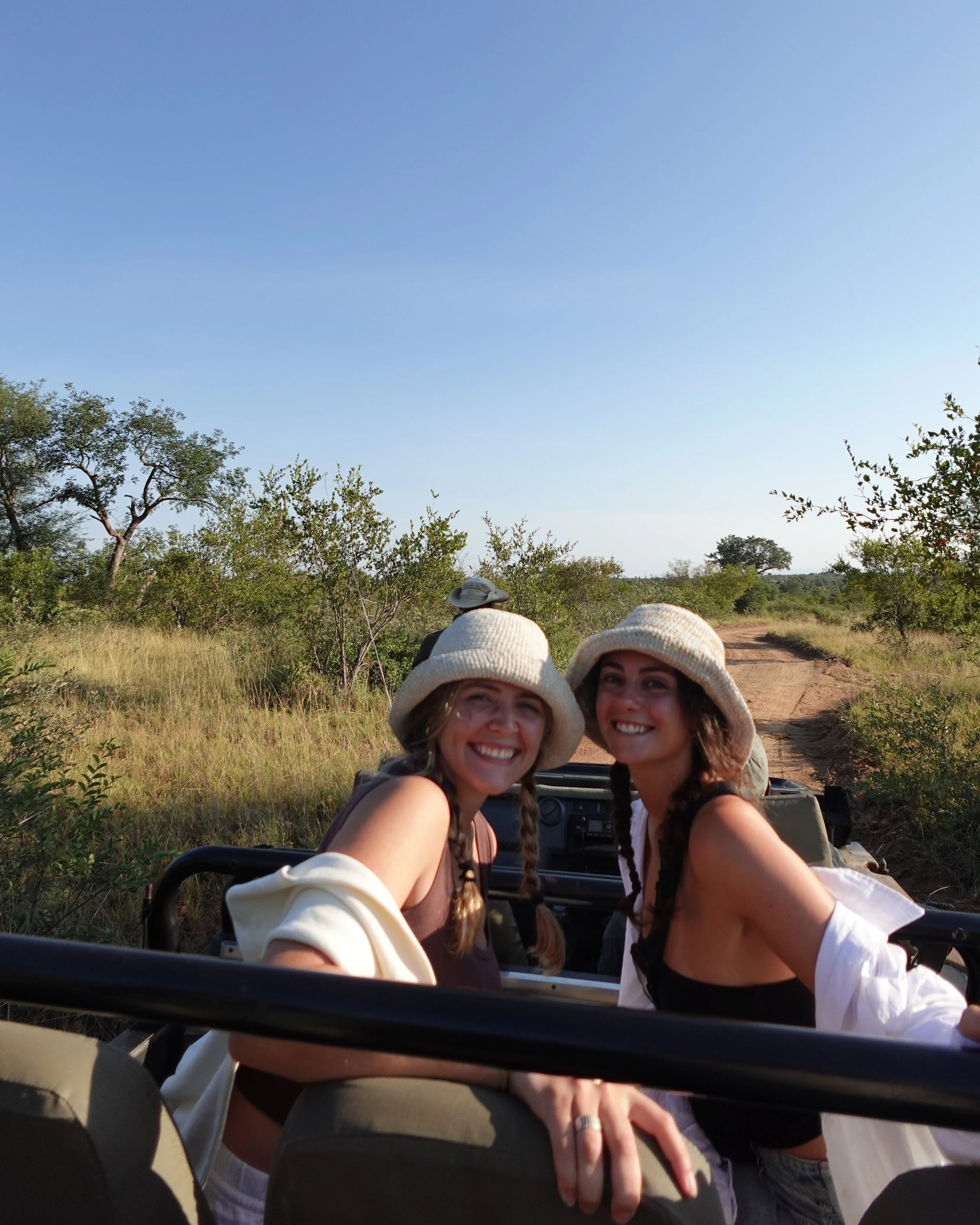 Two women smiling and wearing hats in a safari vehicle on a dirt road in a grassy, wooded area under a clear blue sky.