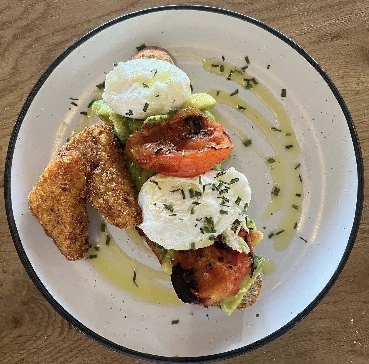 Open-faced sandwich with avocado, grilled tomatoes, poached eggs, and fried fish on a white plate with a black rim
