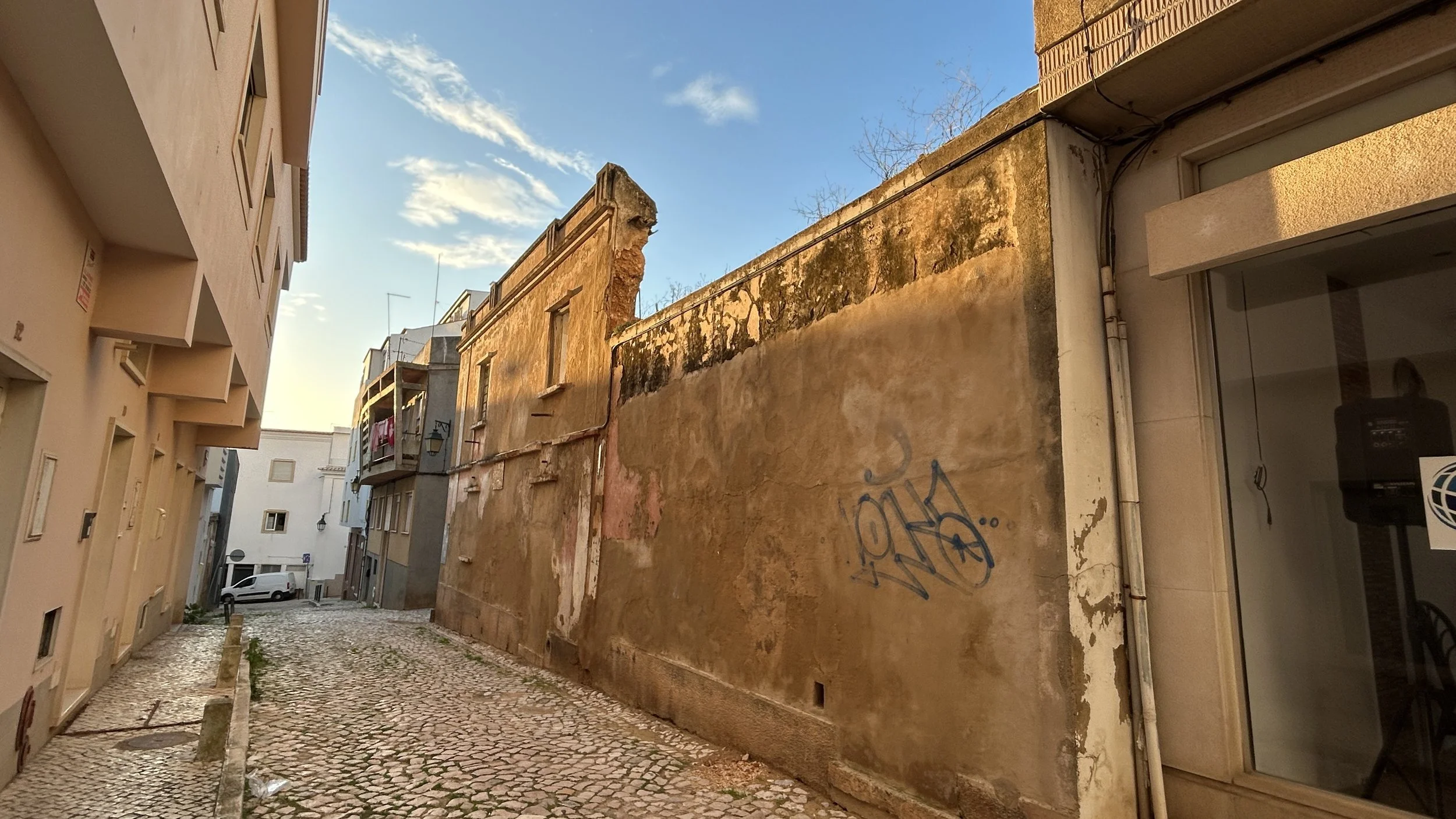 Sunlit narrow cobblestone street between modern and old buildings, with peeling walls and graffiti, under a blue sky with clouds in the centre of Portimão.