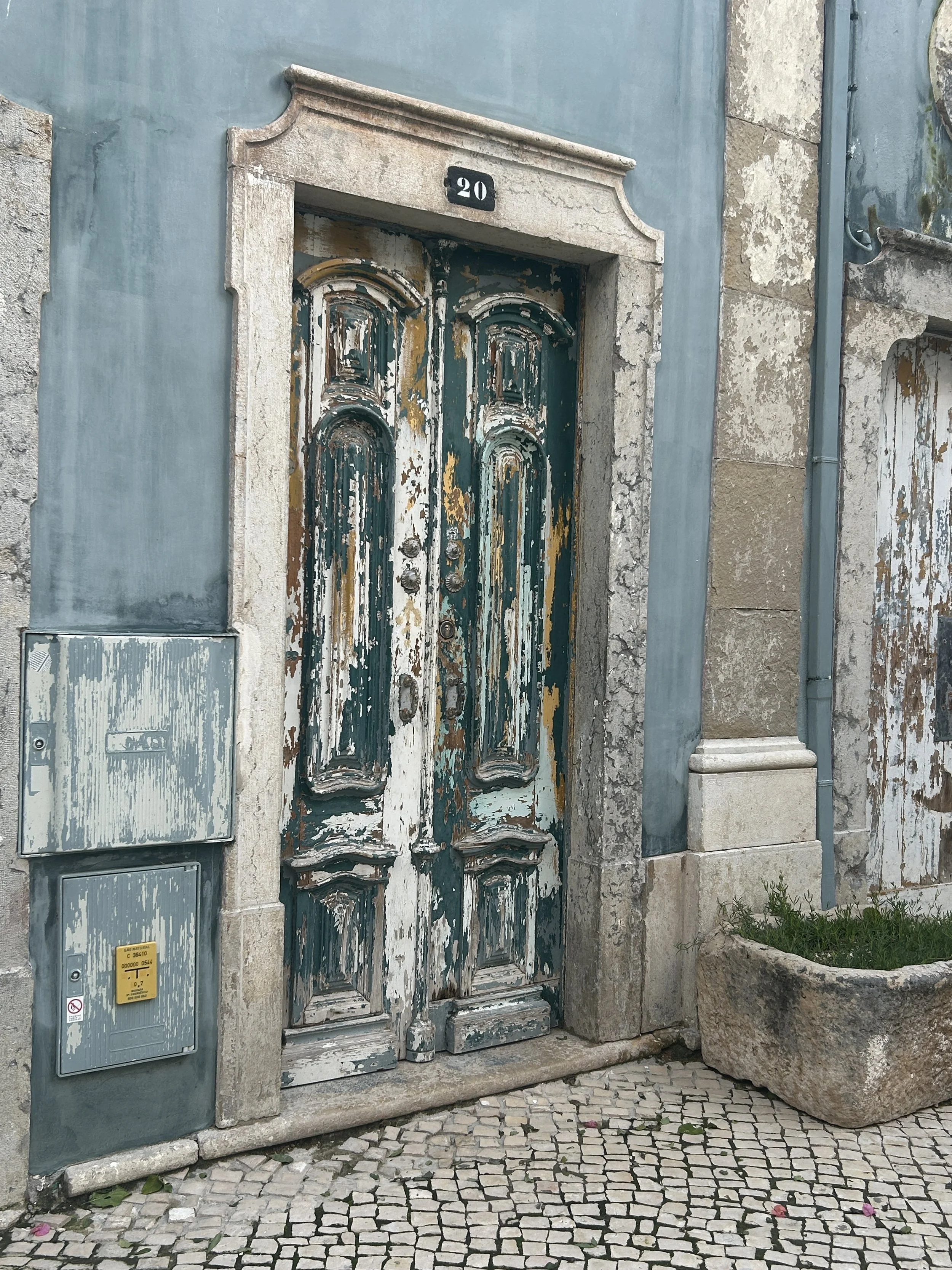 Old, weathered Portuguese wooden door with peeling paint, set in a stone frame on a building numbered 20, with a cobblestone sidewalk in front and a large stone planter to the right.