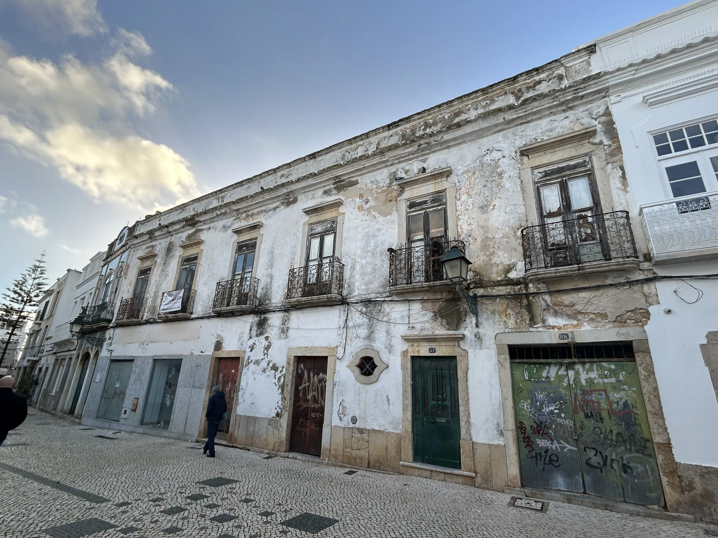 Old, weathered building with peeling paint, iron balconies, and graffiti on metal doors, located on a cobblestone street under a partly cloudy sky.