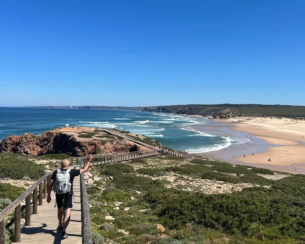 A person walking on a wooden boardwalk towards a beach, with ocean waves, sandy shoreline, and cliffs in the background on a sunny day.
