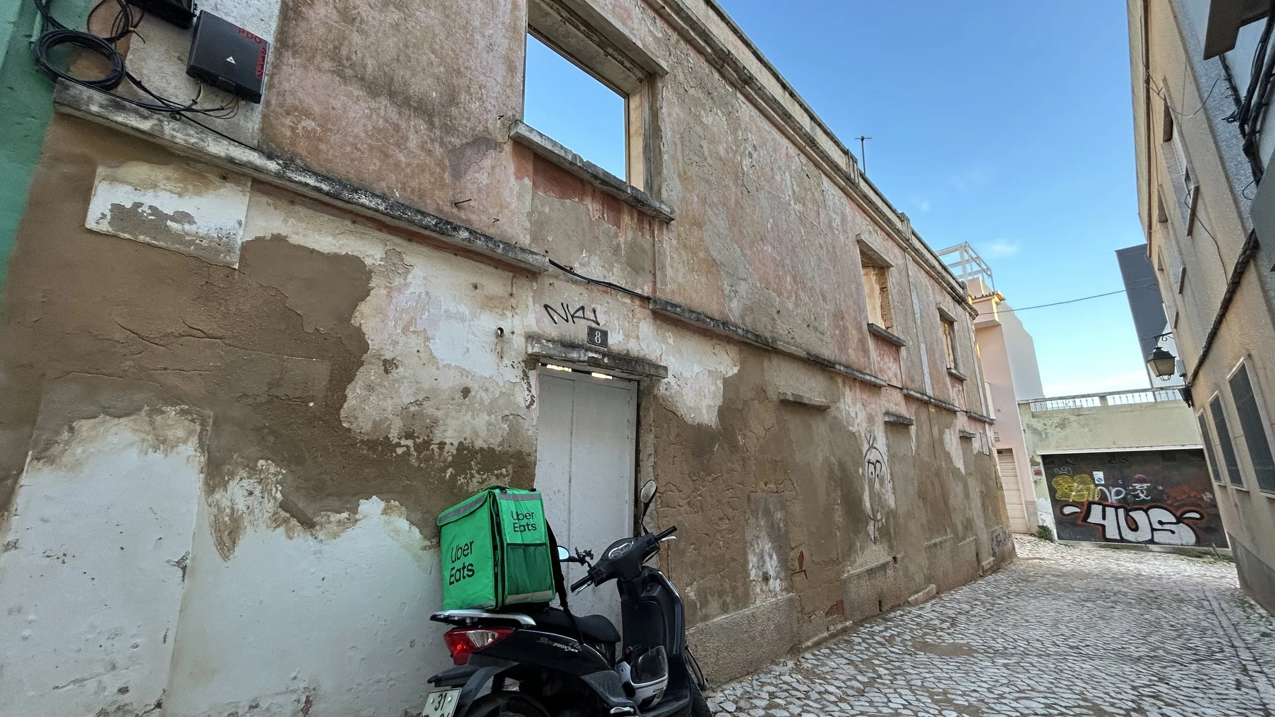 A black scooter with an Uber Eats delivery bag parked on a cobblestone street next to a weathered, peeling wall with graffiti in the background, under a clear blue sky in the centre of Portimão.