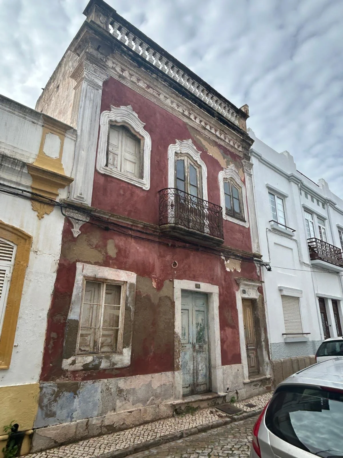 An old, weathered three-story building with a red facade, peeling paint, and wooden shutters in a historic neighborhood, with neighboring buildings and parked cars.