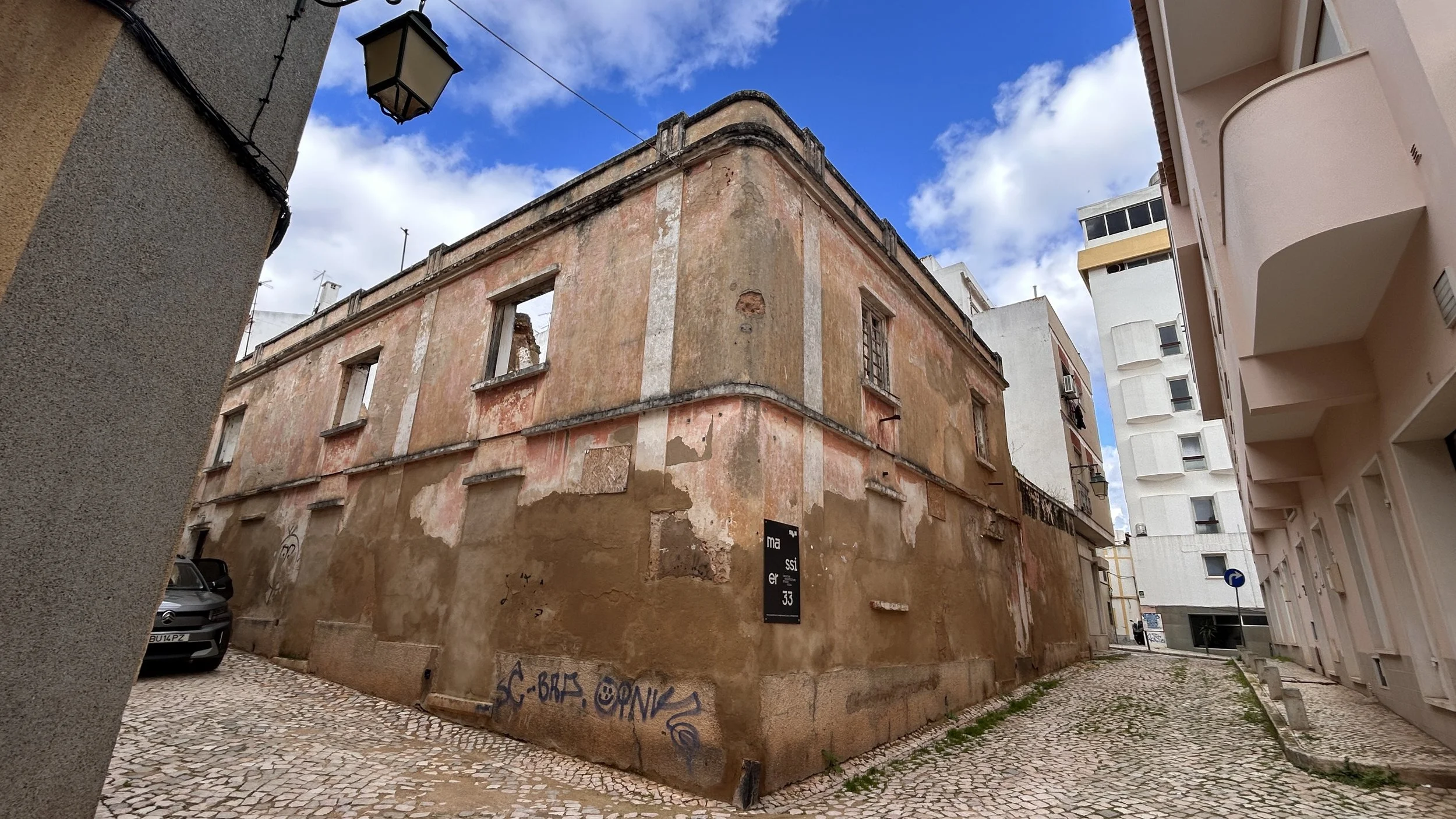 Old, weathered building with peeling paint and boarded-up windows on a corner, surrounded by newer buildings, cobblestone street, and a partly cloudy blue sky.