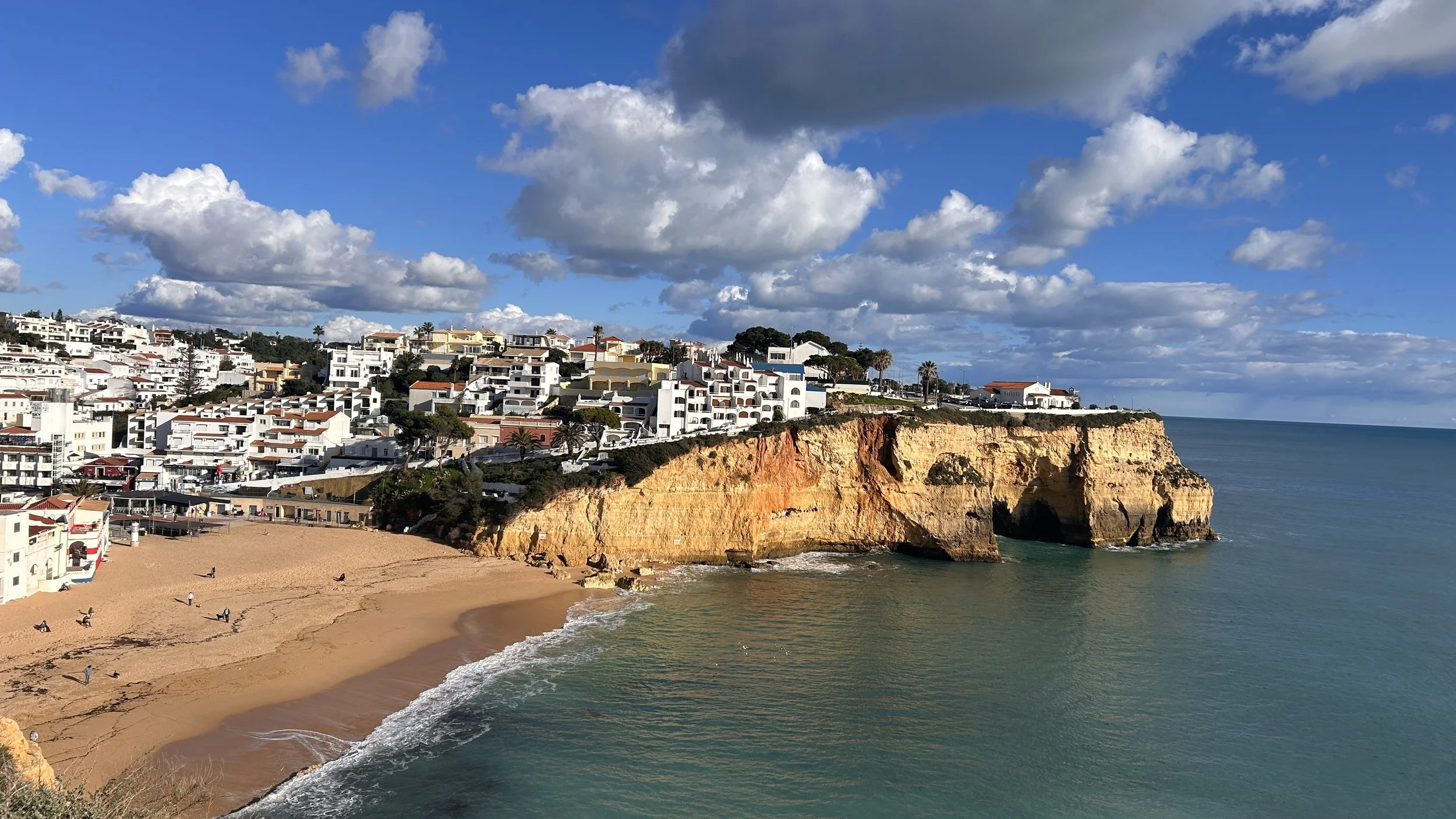 Clifftop view of a sandy beach with white residential buildings on a hill, ocean waves, and a partly cloudy sky.