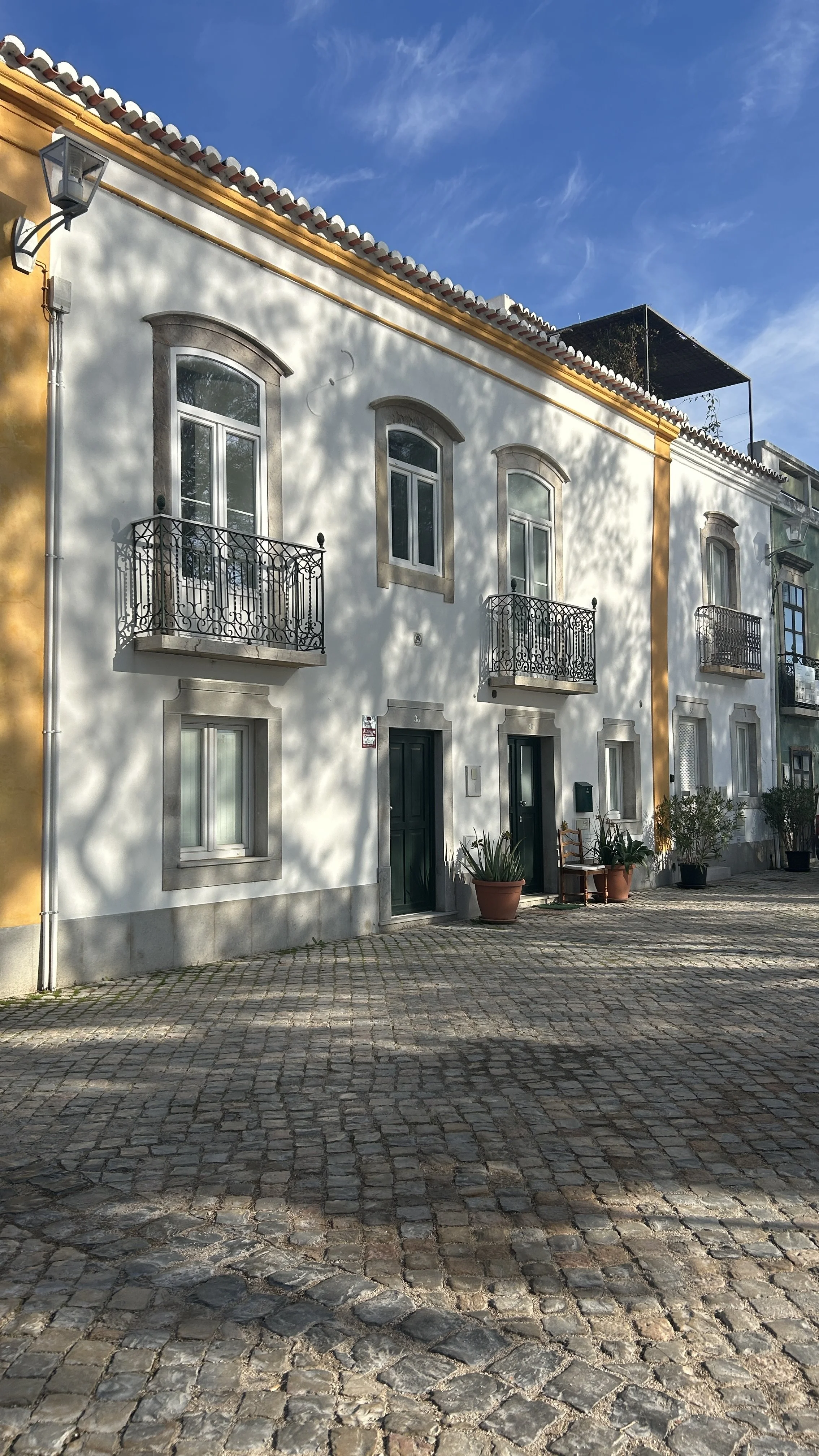 A white building with three black balconies on the second floor, arched windows, and a cobblestone street in front. There are potted plants outside and shadows of tree branches on the building. The sky is blue with some clouds.