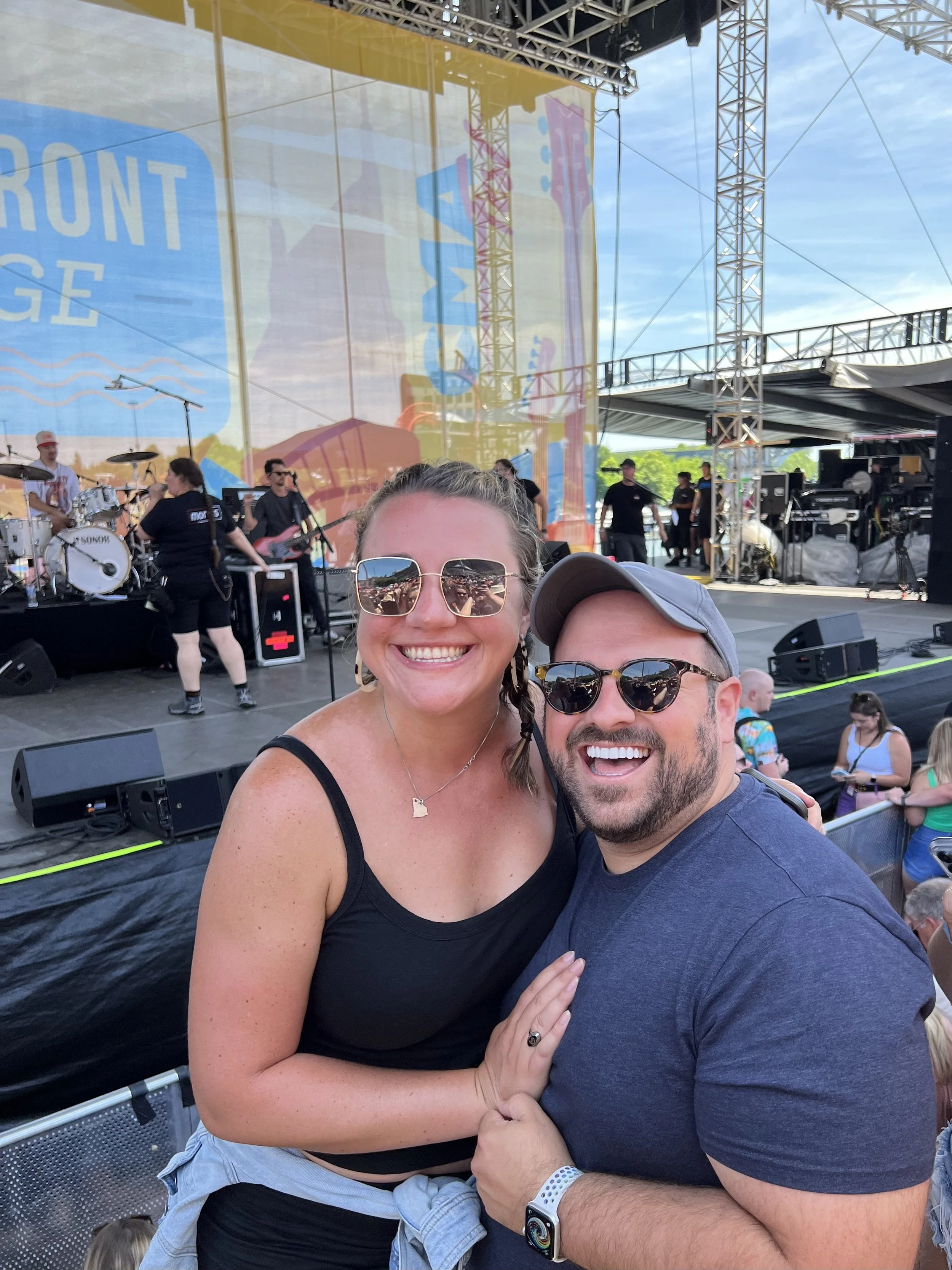 A smiling couple at an outdoor music festival or concert, standing close together, with a live band performing on a stage behind them and a large colorful screen in the background.