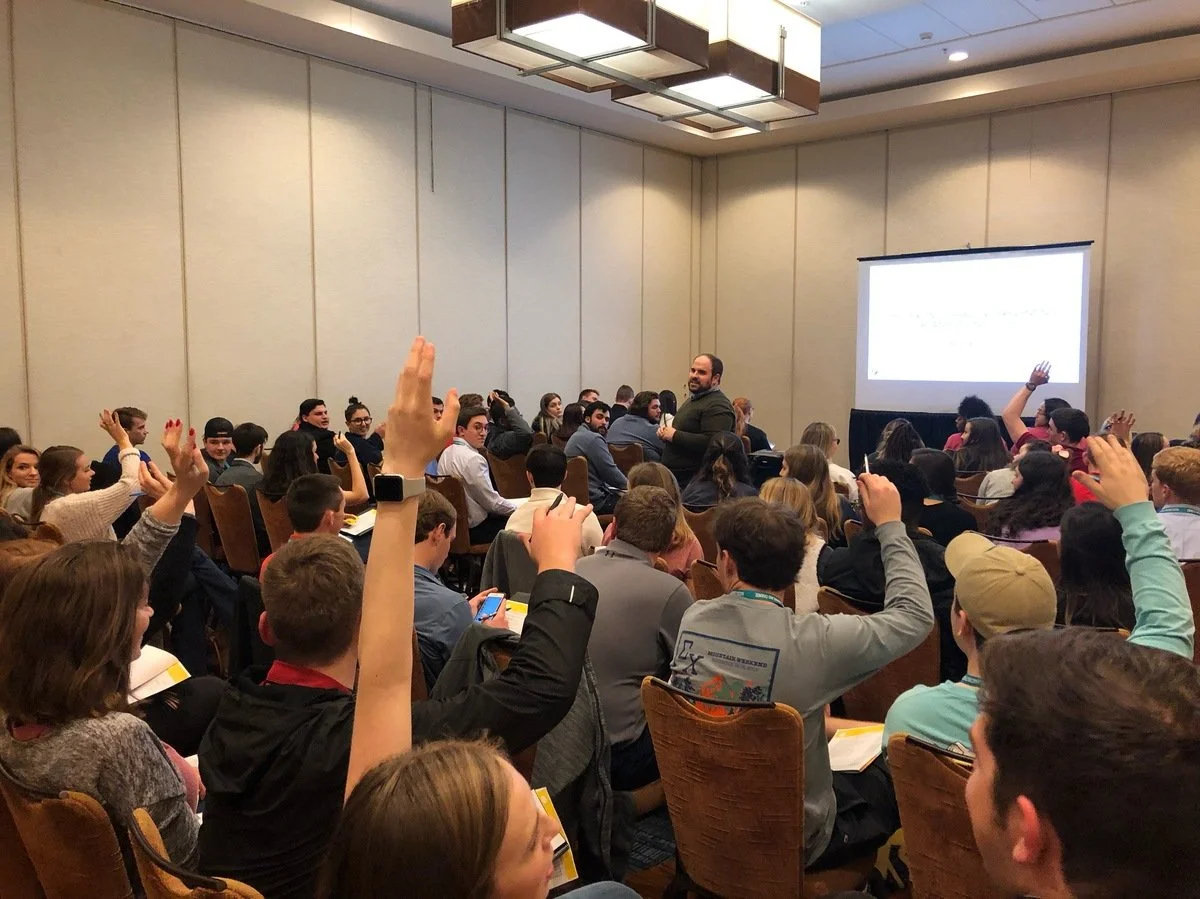 A conference room filled with people, many of whom are raising their hands. A speaker stands near a screen at the front of the room, engaging with the audience.