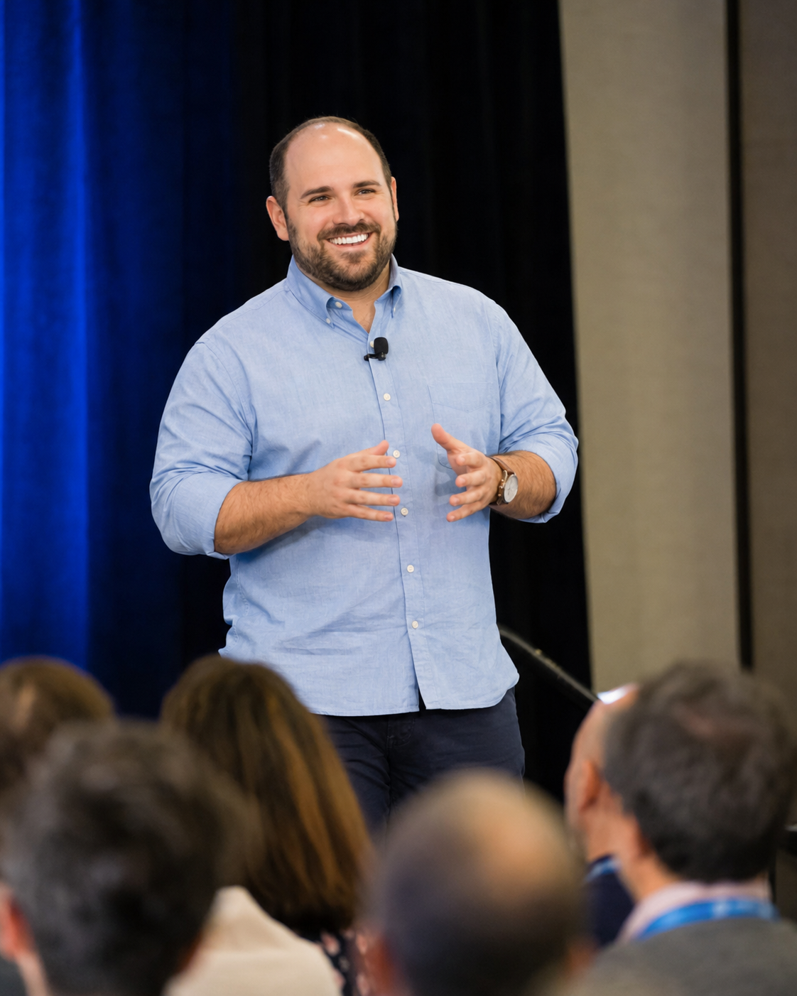A man with a beard and mustache smiling, wearing a light blue shirt and a wristwatch, giving a presentation to an audience in a conference room.