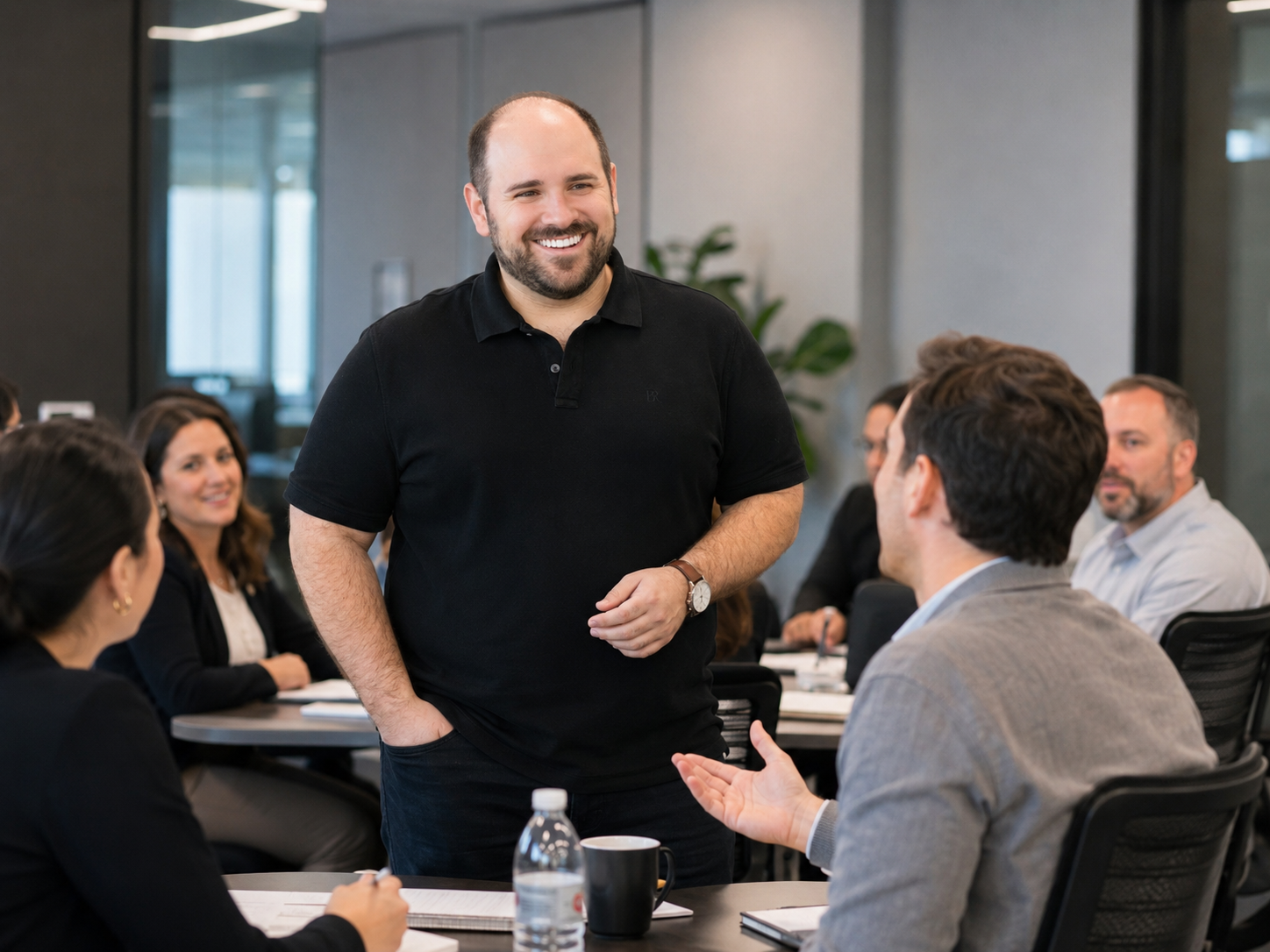 A smiling man in a black polo shirt standing and talking to a group of people seated around a conference table in a modern office.