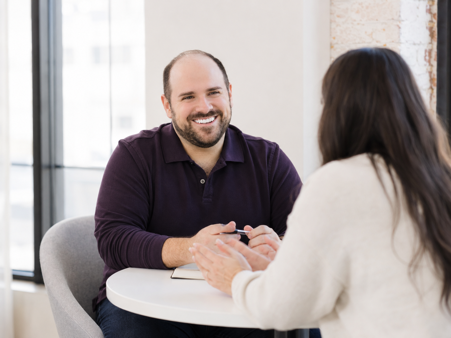 A man and a woman sitting at a round table having a conversation in a modern office space. The man is smiling and holding a pen, while the woman has her hands on the table and is facing away from the camera.