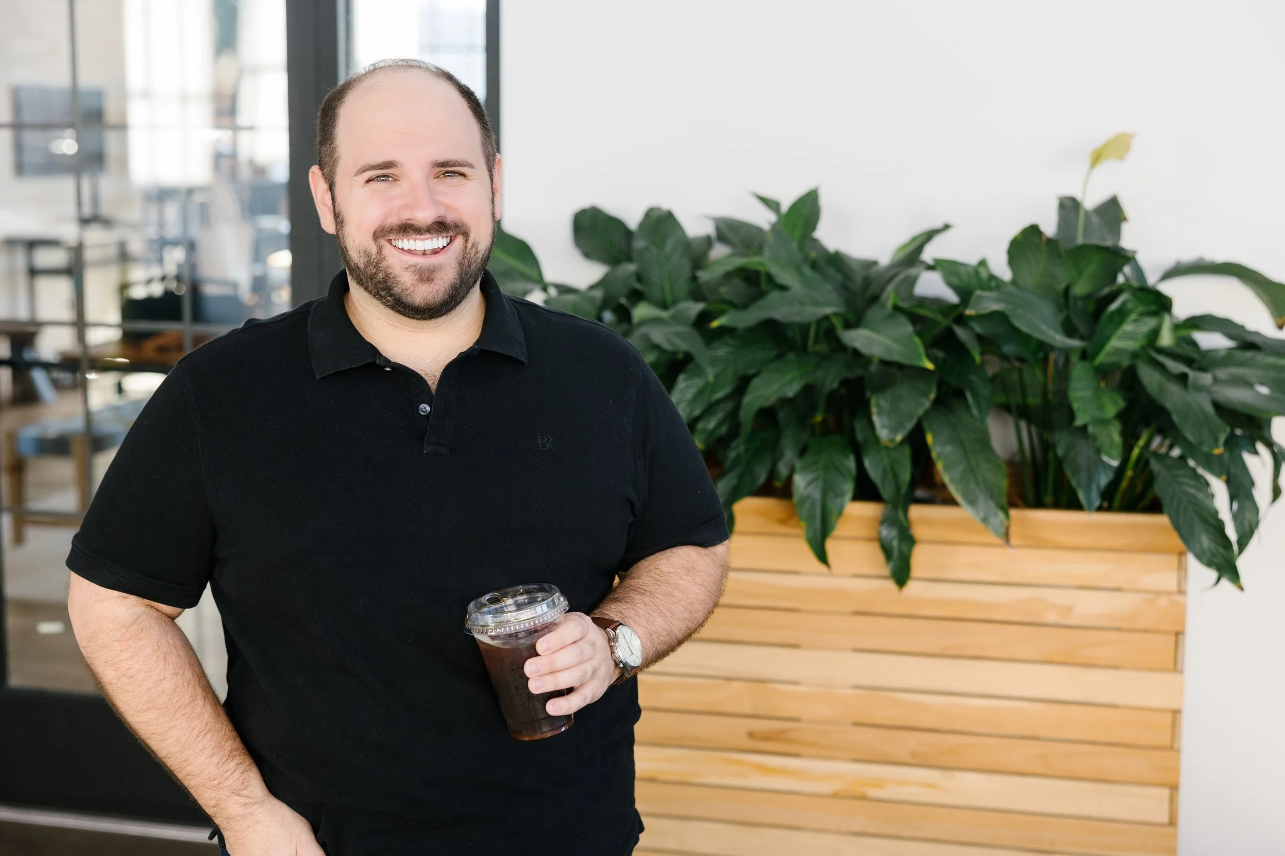 A man with a beard and short hair smiling while holding a cup of iced coffee in an indoor setting with green plants and wooden furniture.