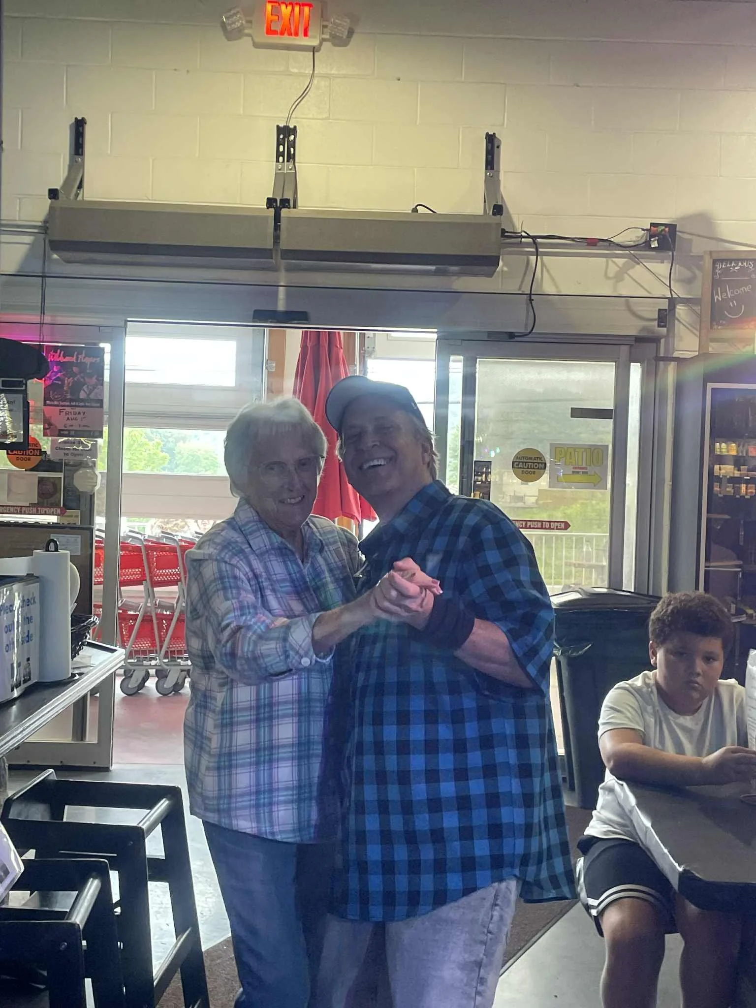 A smiling elderly woman and a smiling man dancing together inside a restaurant or cafe. A young boy sits at a table nearby. The background shows a door with glass windows, signs, and red chairs outside.