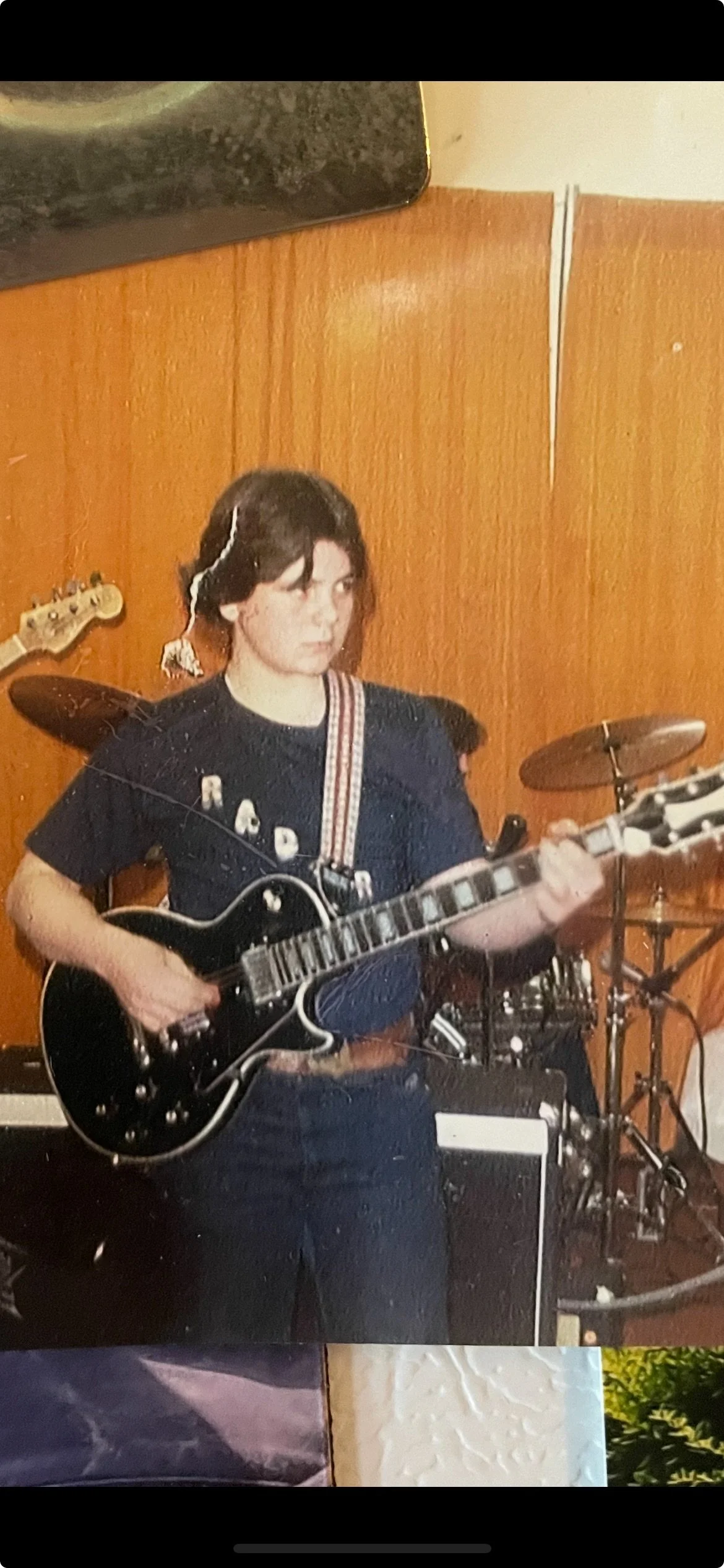 A woman with shoulder-length dark hair playing a black electric guitar in a room with wood-paneled walls, wearing a black t-shirt and a striped guitar strap, with drums and part of a bass guitar in the background.