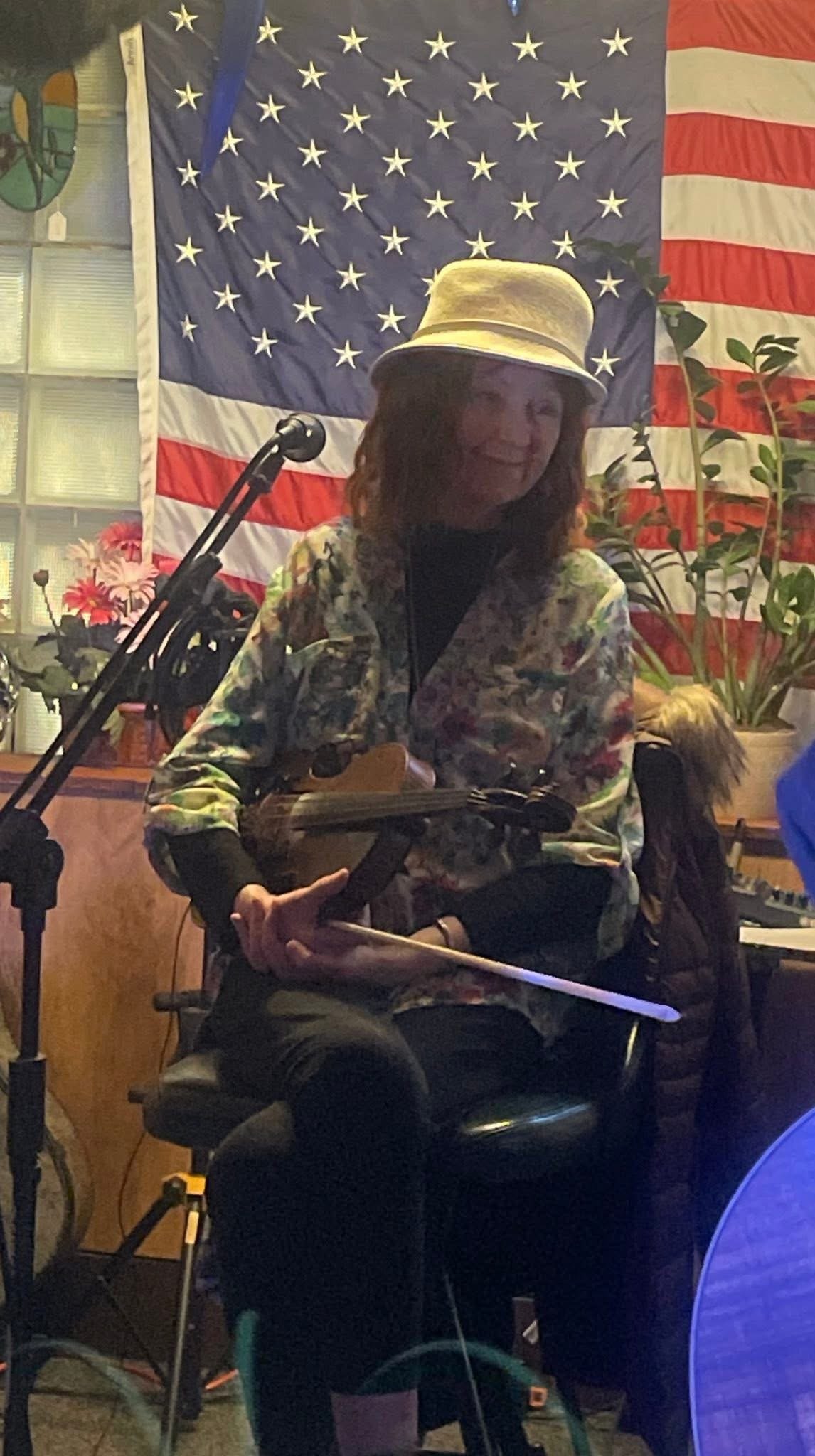 A woman sitting on a chair playing a small guitar or ukulele, wearing a hat and a floral jacket, with an American flag and plants in the background.