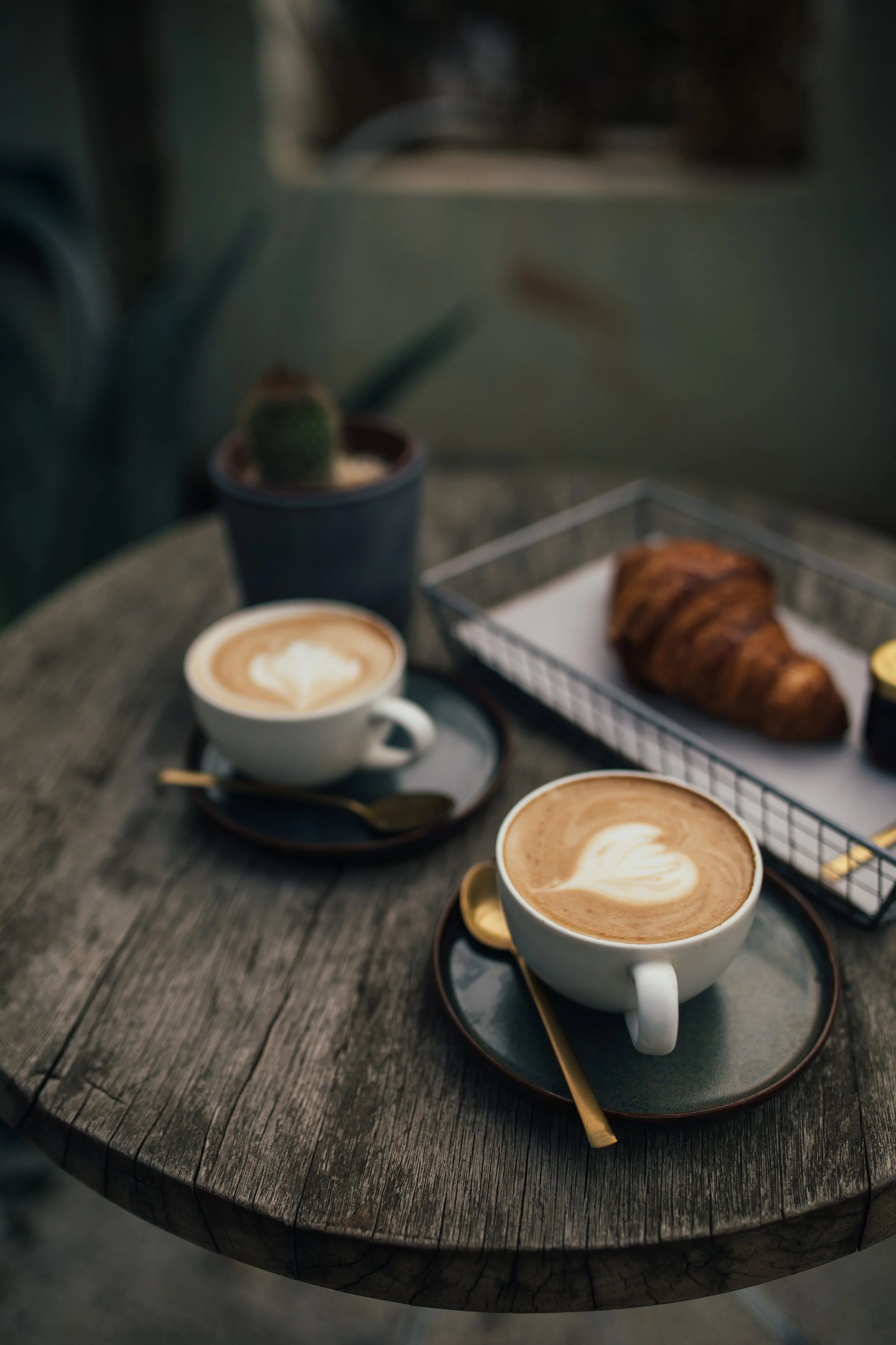 Two cups of coffee with latte art, a croissant on a tray, a small plant in a pot, and a granite container on a wooden table.