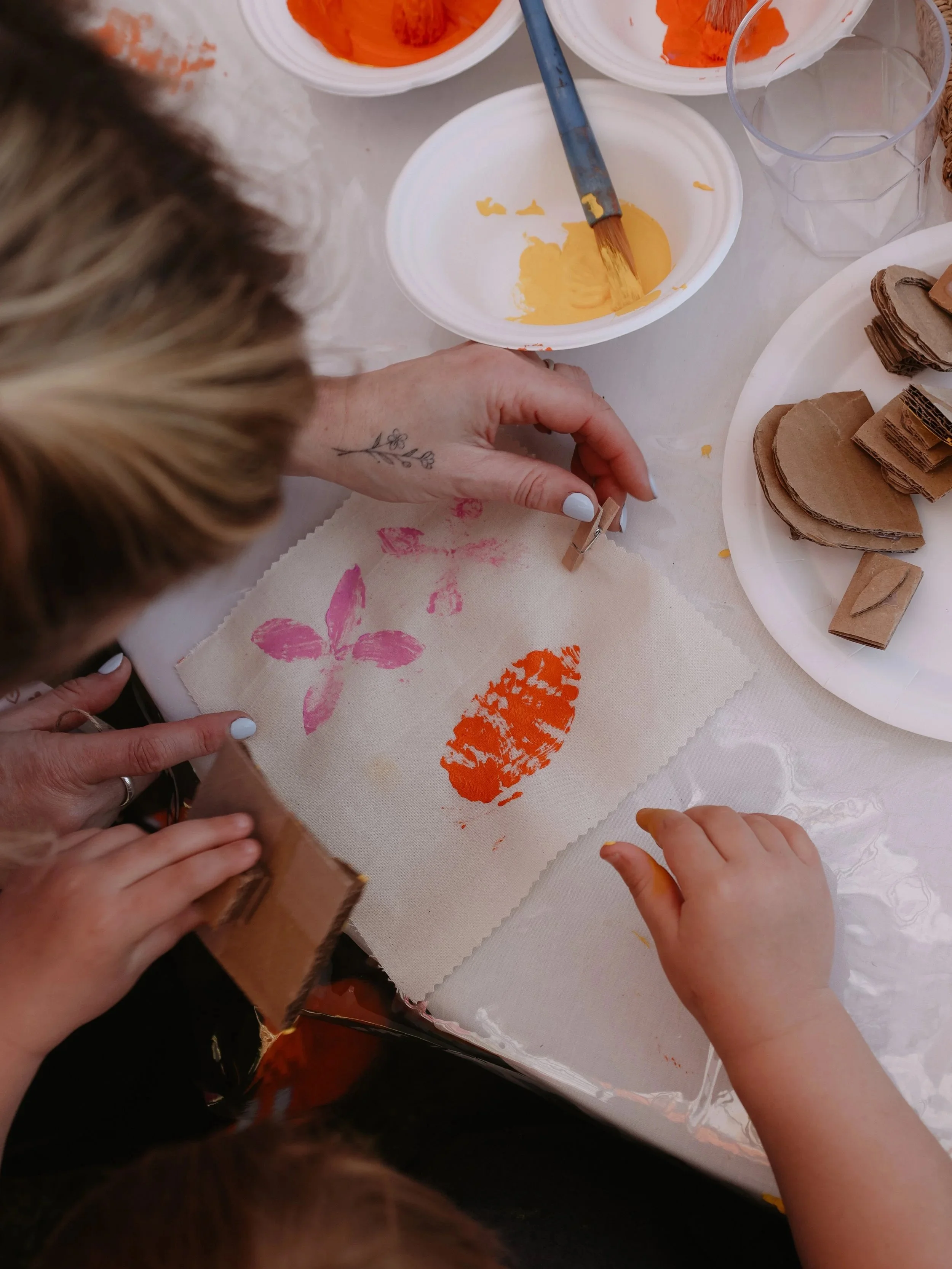 A person and a child are stamping flower and leaf shapes onto fabric with rubber stamps dipped in orange and pink paint. There are bowls of paint, a glass of water, and a plate with cut-outs of cardboard on the table.