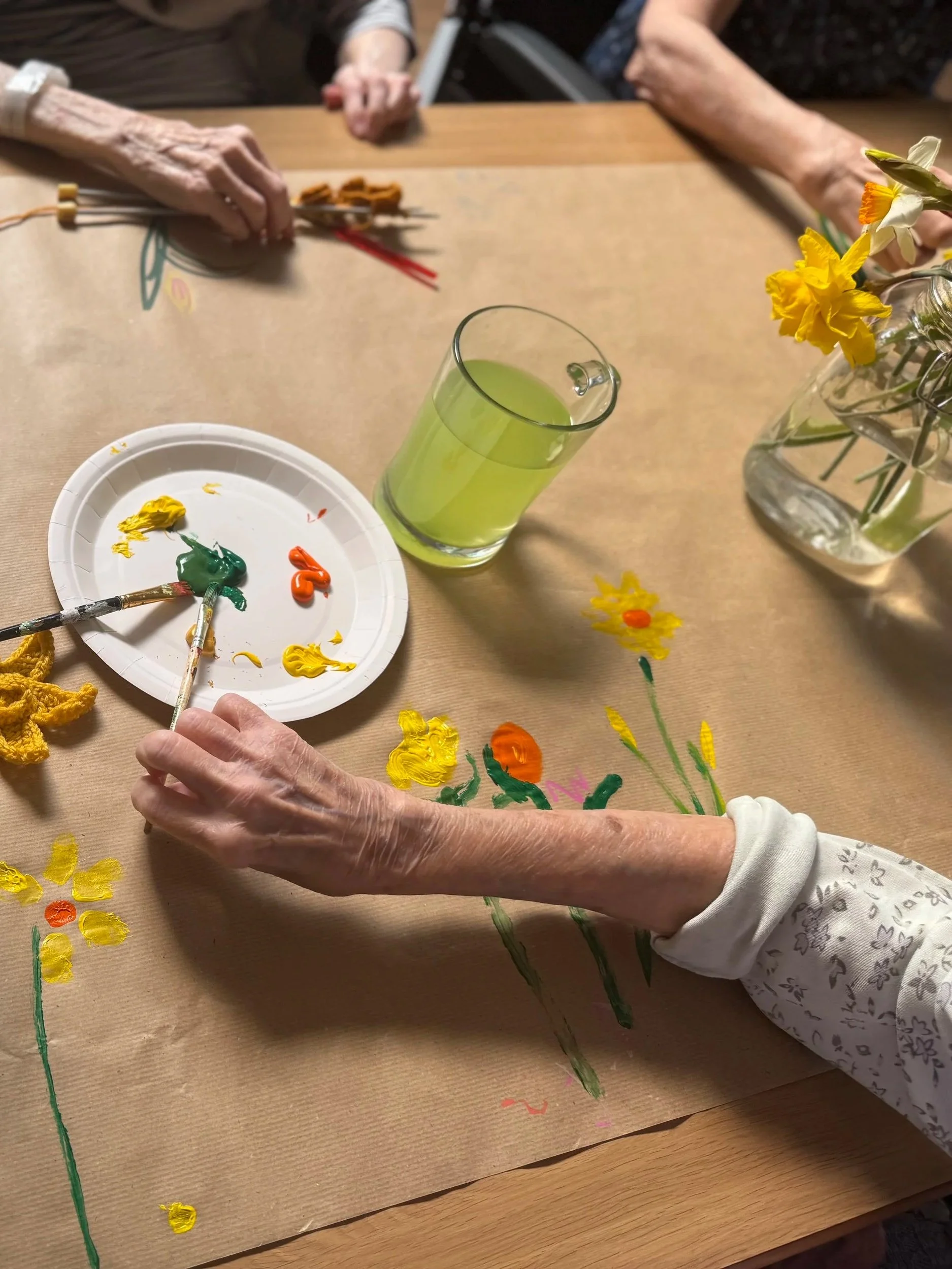 Elderly people painting colorful flowers on a brown paper table with brushes and paints, with a vase of flowers and a glass of green liquid nearby.
