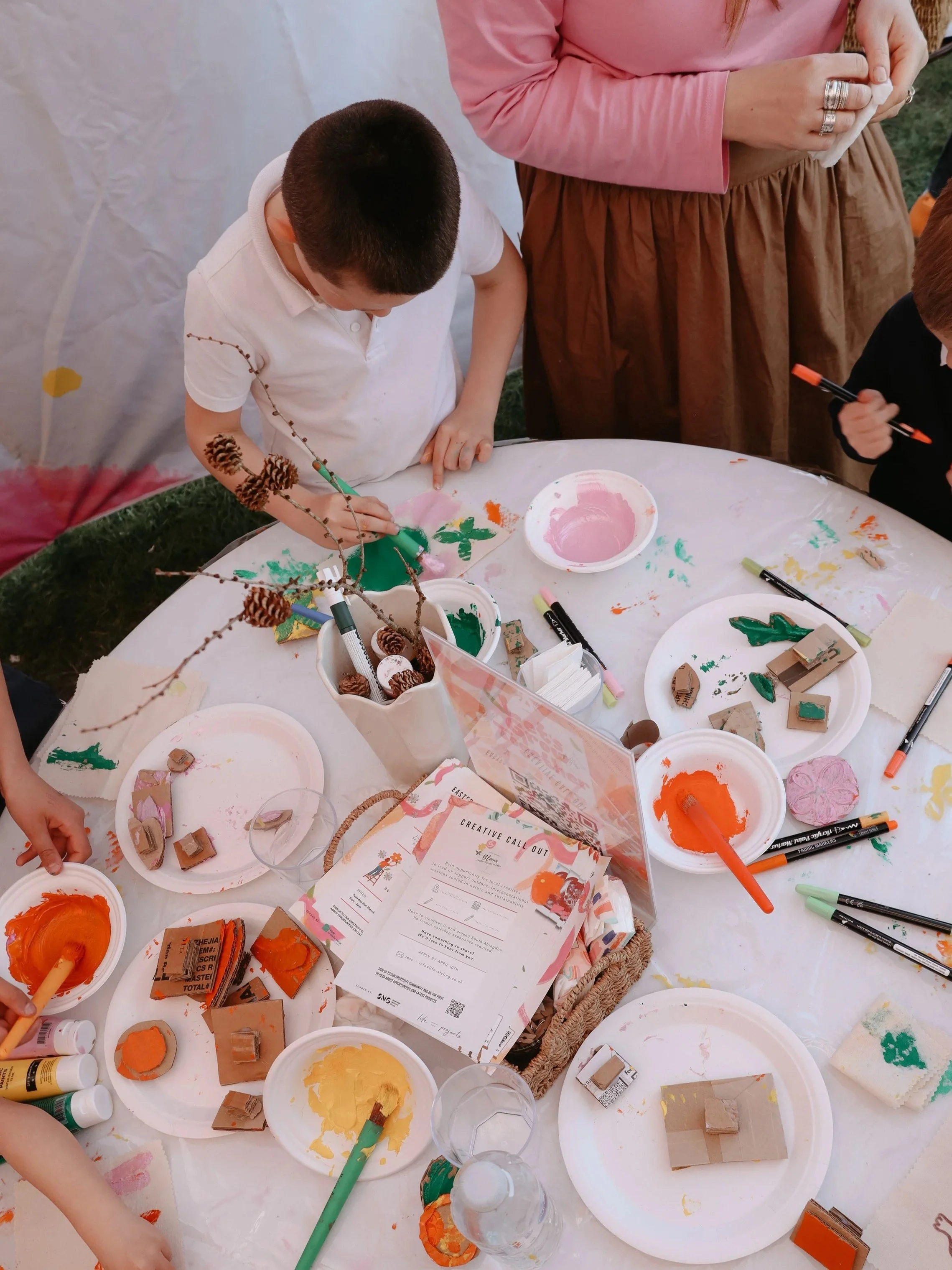 Children at a craft table painting and stamping with various colors and stamps during an outdoor event.