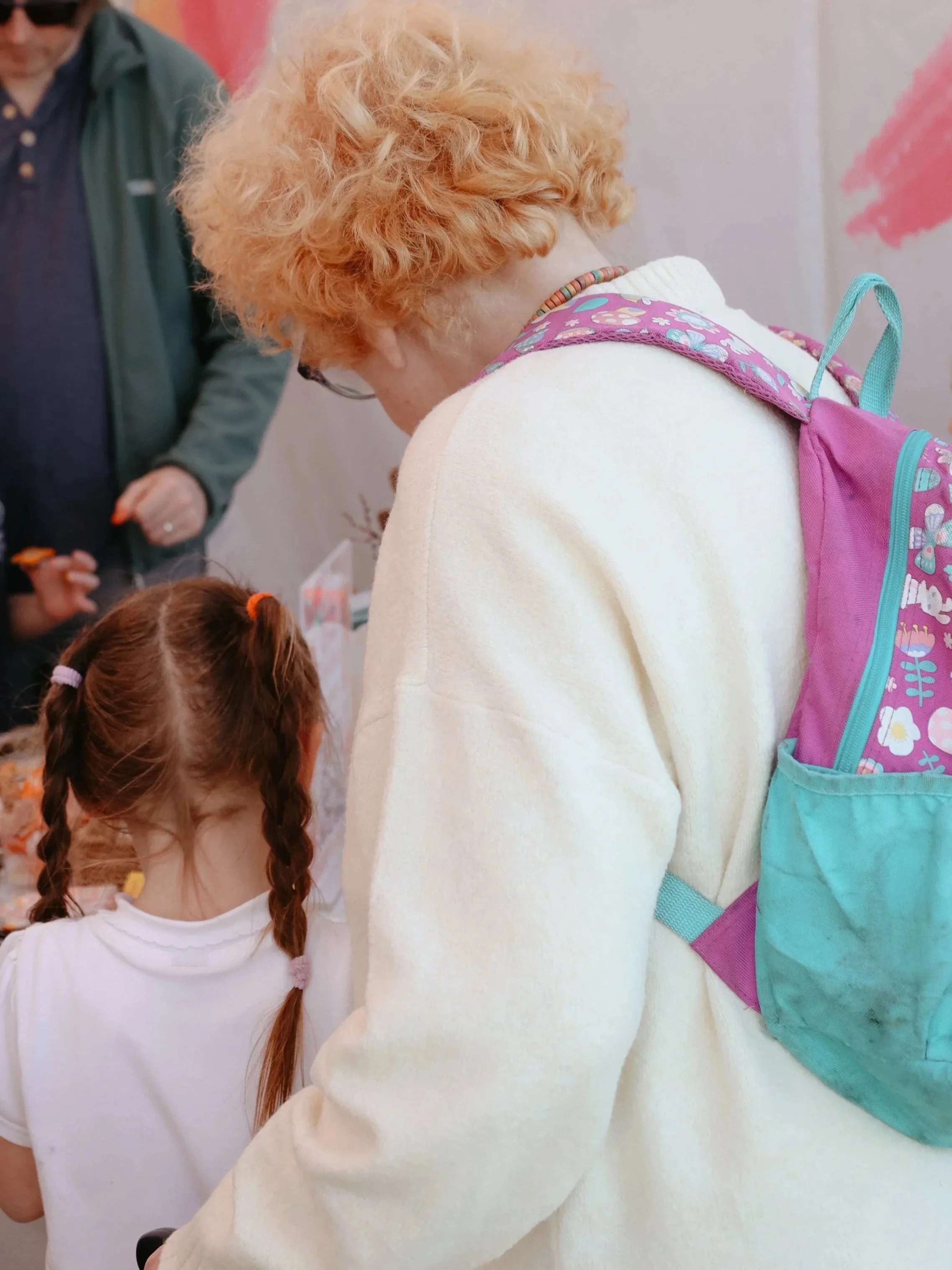A woman with curly red hair and a colorful backpack leaning towards a young girl with braided hair in a white shirt at an indoor event with pink and orange decorations. Intergenerational creativity Abingdon